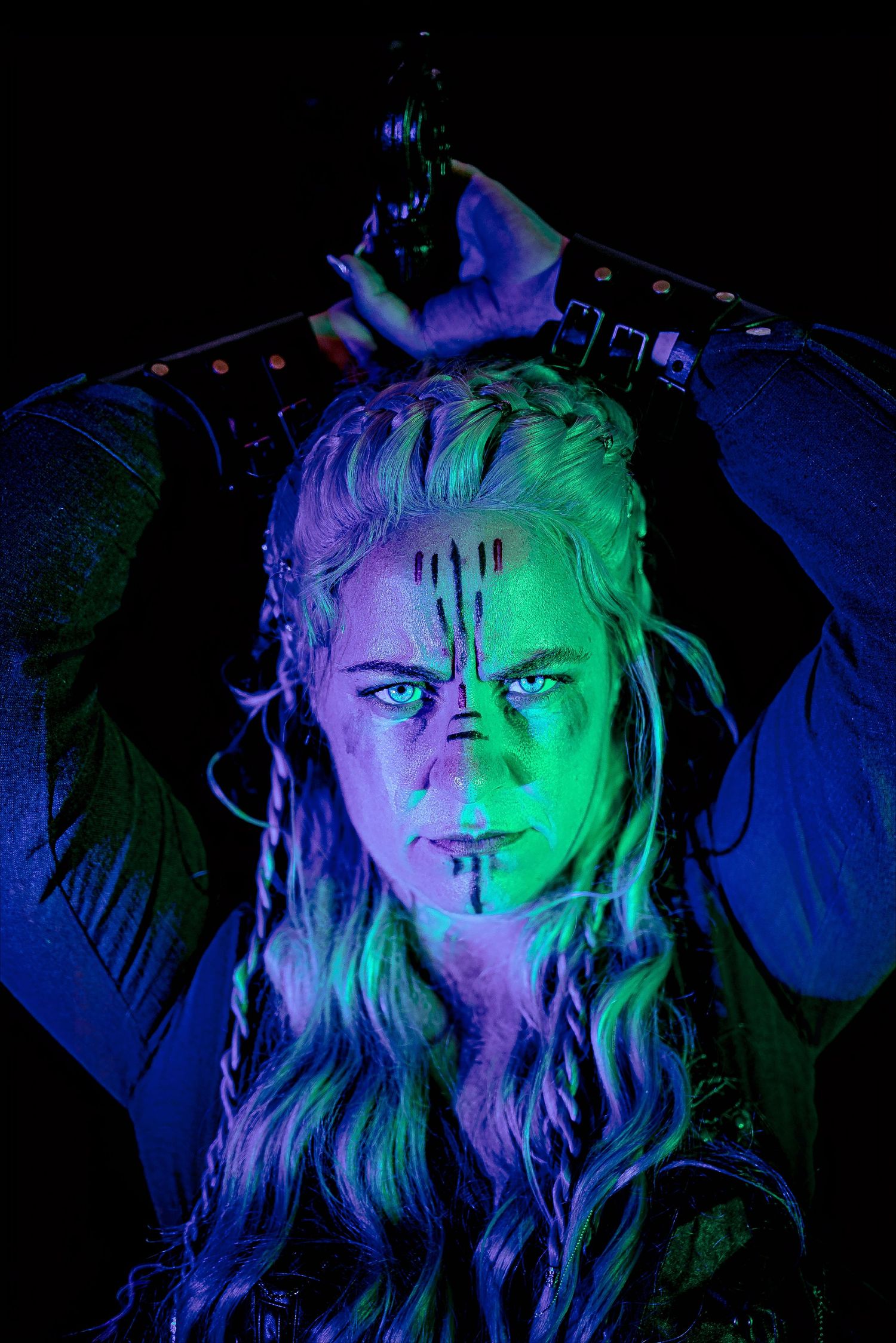 A fierce warrior woman with braided hair, glowing blue-green UV lighting, dark face paint streaks, in dramatic fantasy costume.
