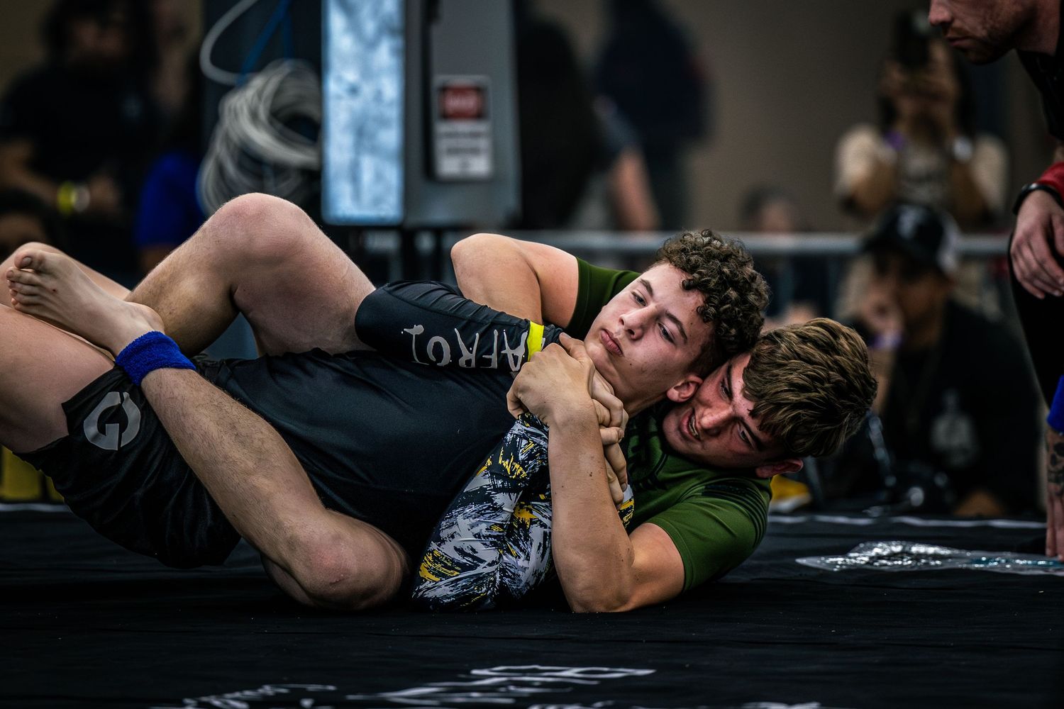 Two young grapplers compete in a Brazilian jiu-jitsu match on a black mat during an indoor martial arts tournament.