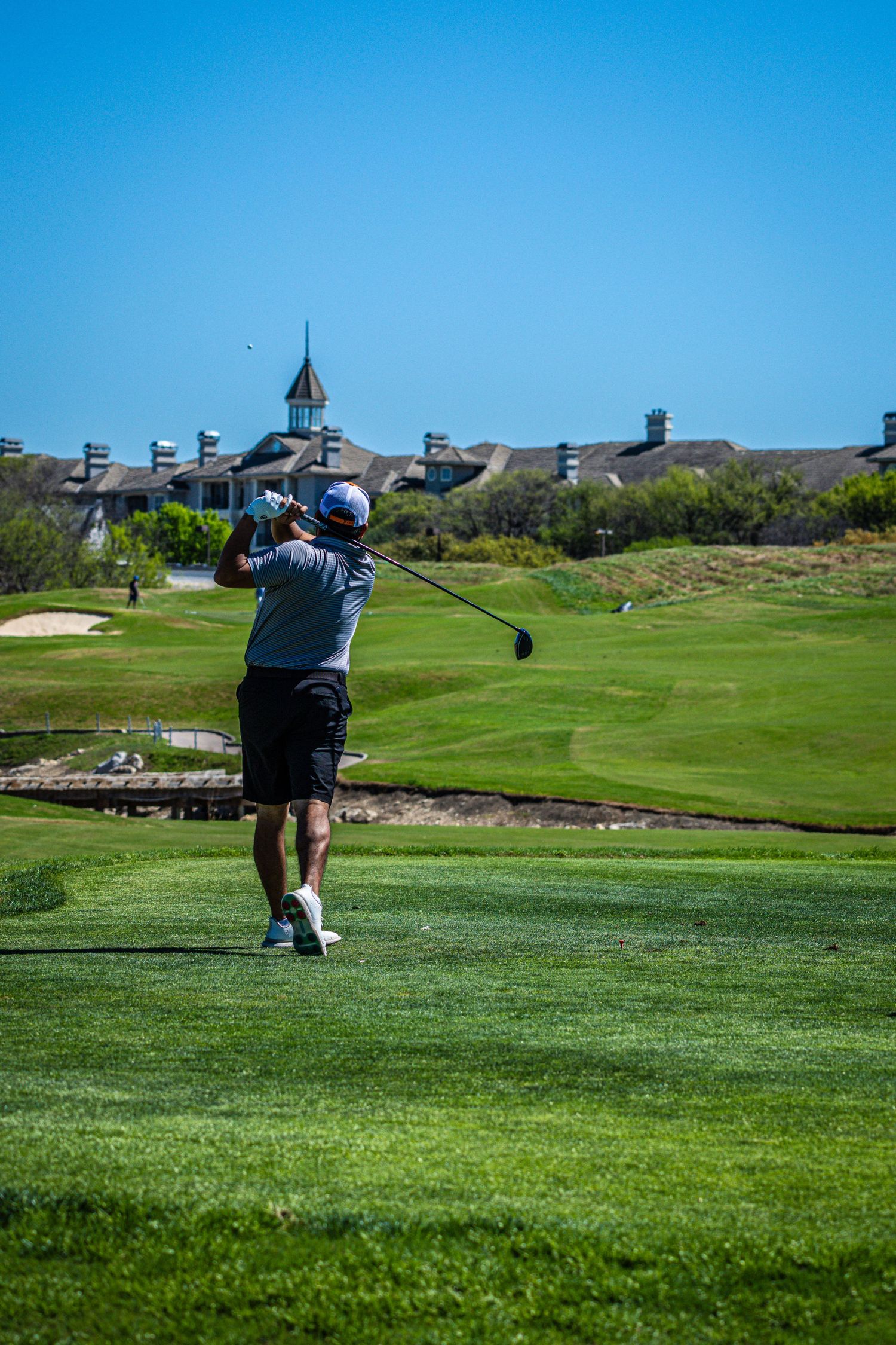 Golfer mid-swing on a lush green golf course with a clubhouse and clear blue sky in the background.