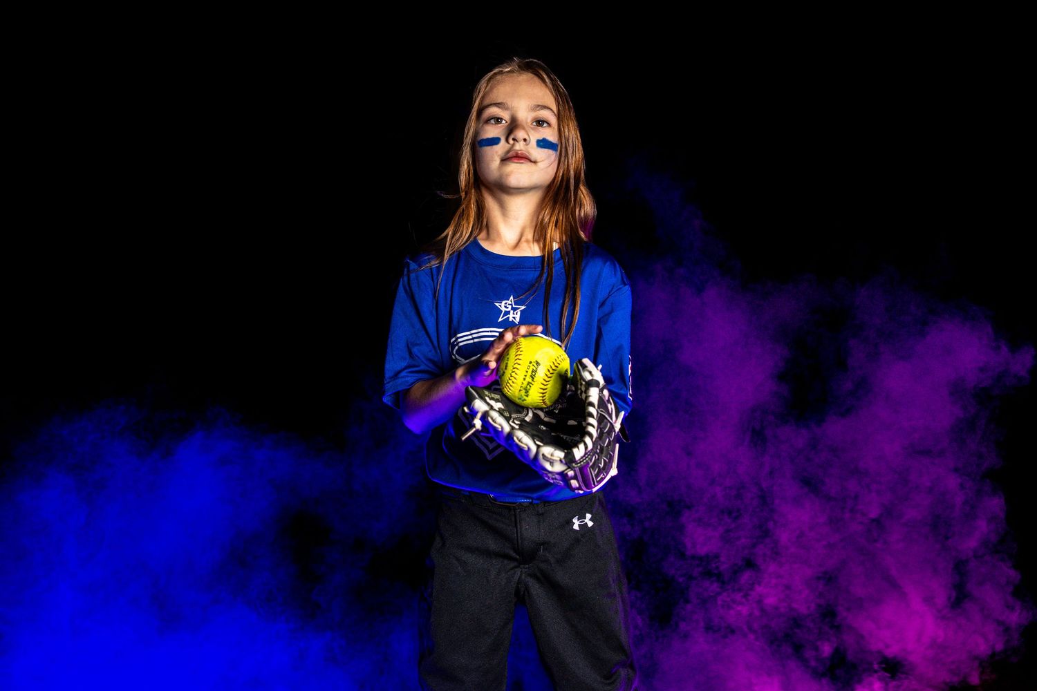 Young girl in blue softball uniform holds yellow helmet with purple and blue smoke clouds on dark background.