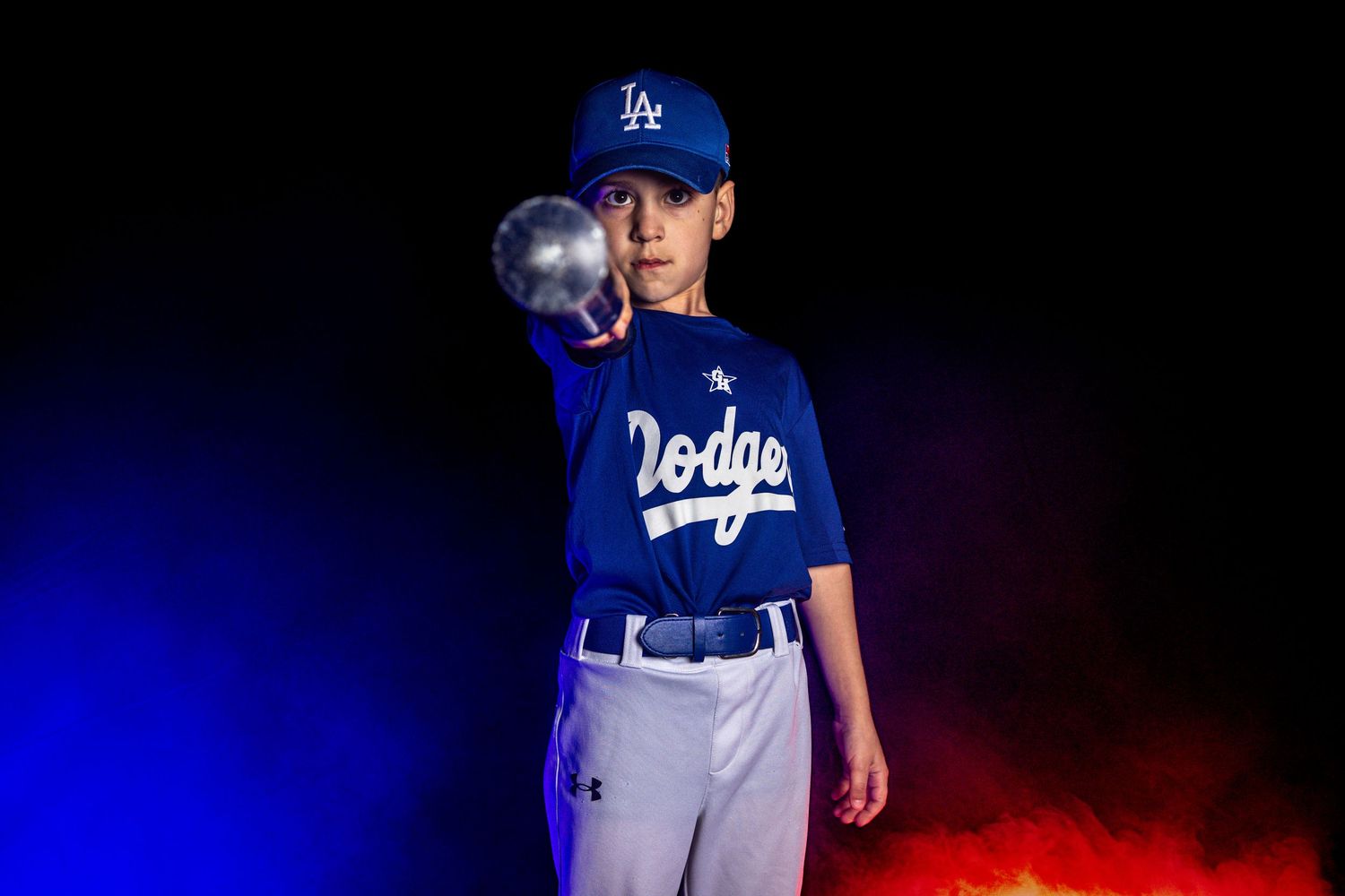 Young boy in Dodgers baseball uniform pitches ball with dramatic red and blue lighting.