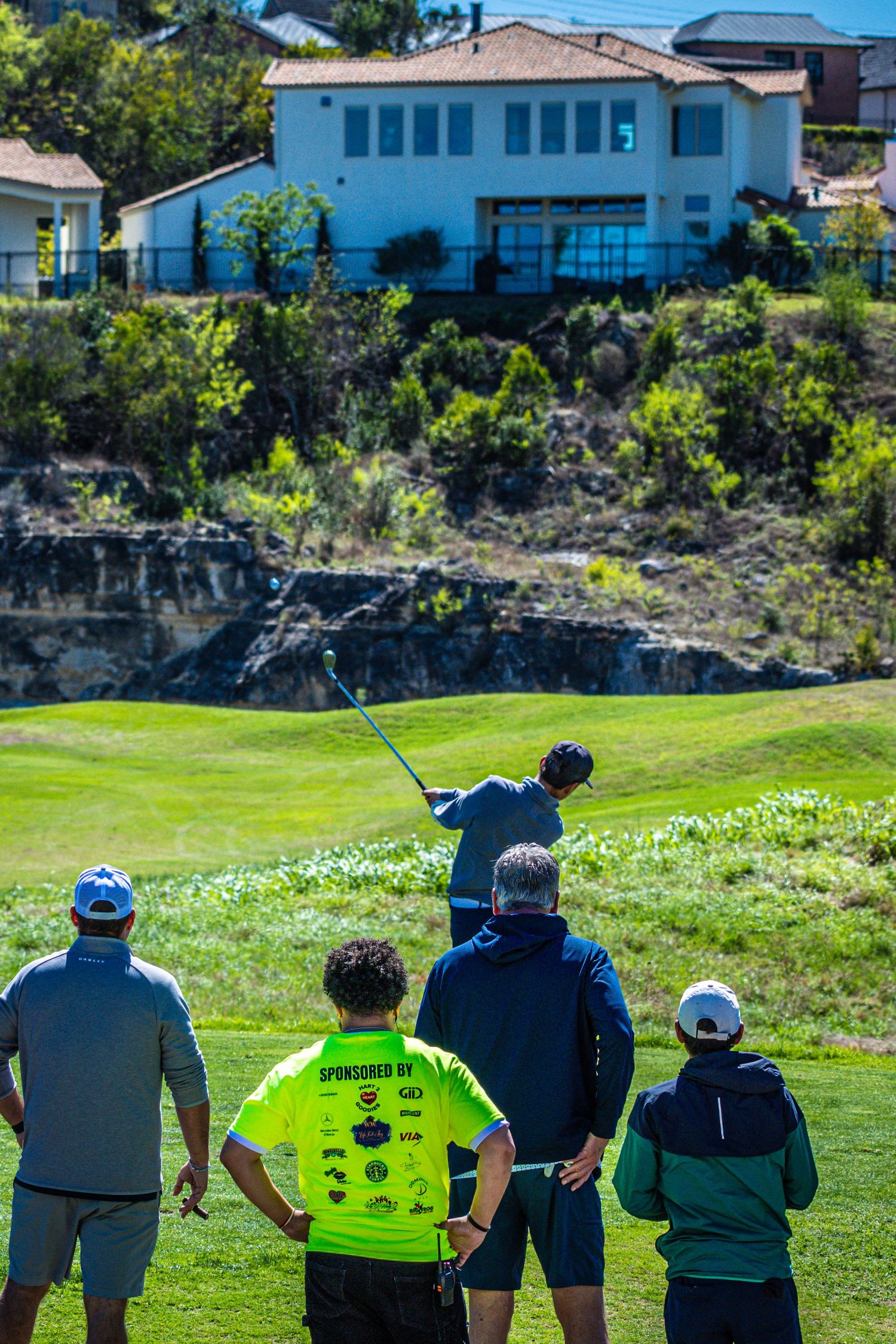 Golfer swings club on lush green course as spectators watch, with a hillside home visible in the background.