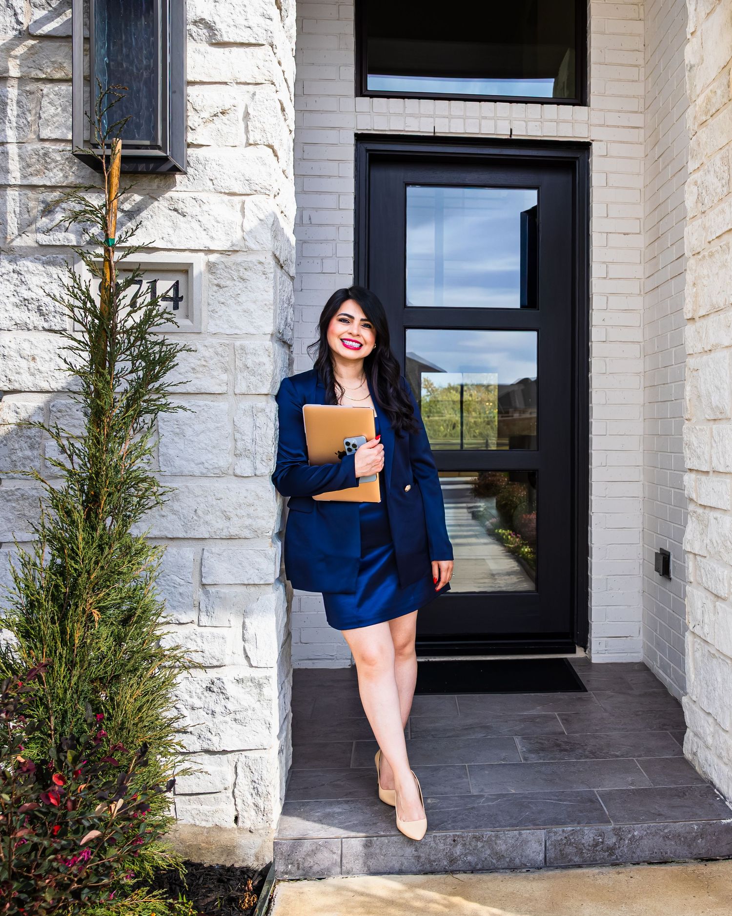 A person in a navy blazer and dress stands confidently in front of modern black doors and white stone exterior.