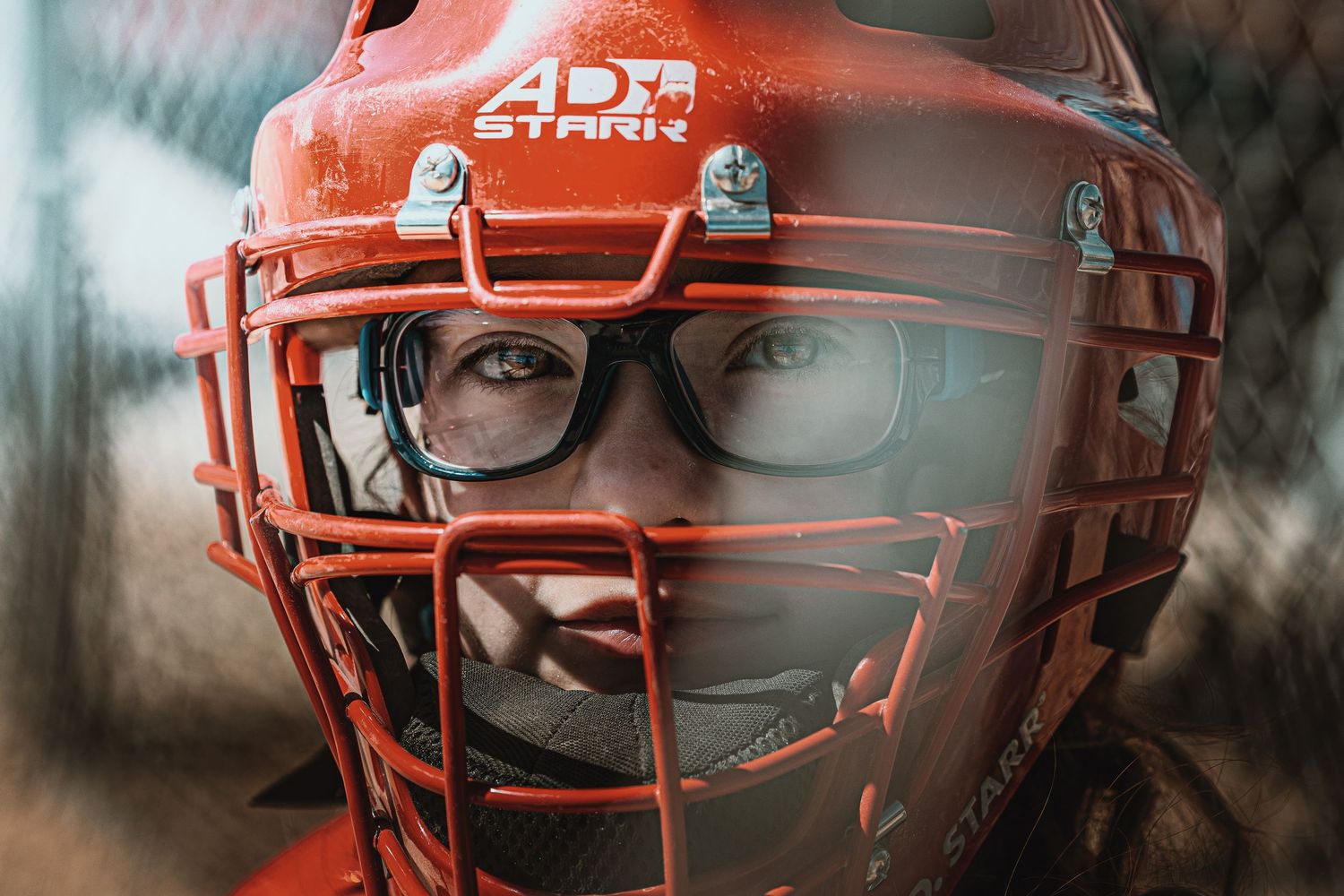 Young baseball catcher wearing red helmet with face guard and glasses, close-up portrait shot.