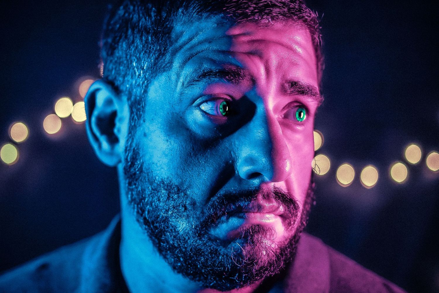 Close-up portrait of a bearded man lit with blue and pink neon lights, looking sideways with a worried expression.