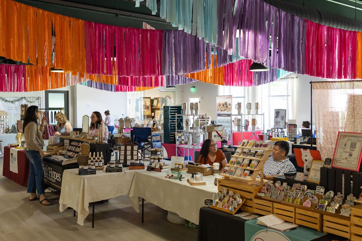 Colorful indoor market with vibrant hanging streamers and vendor tables displaying handmade goods.