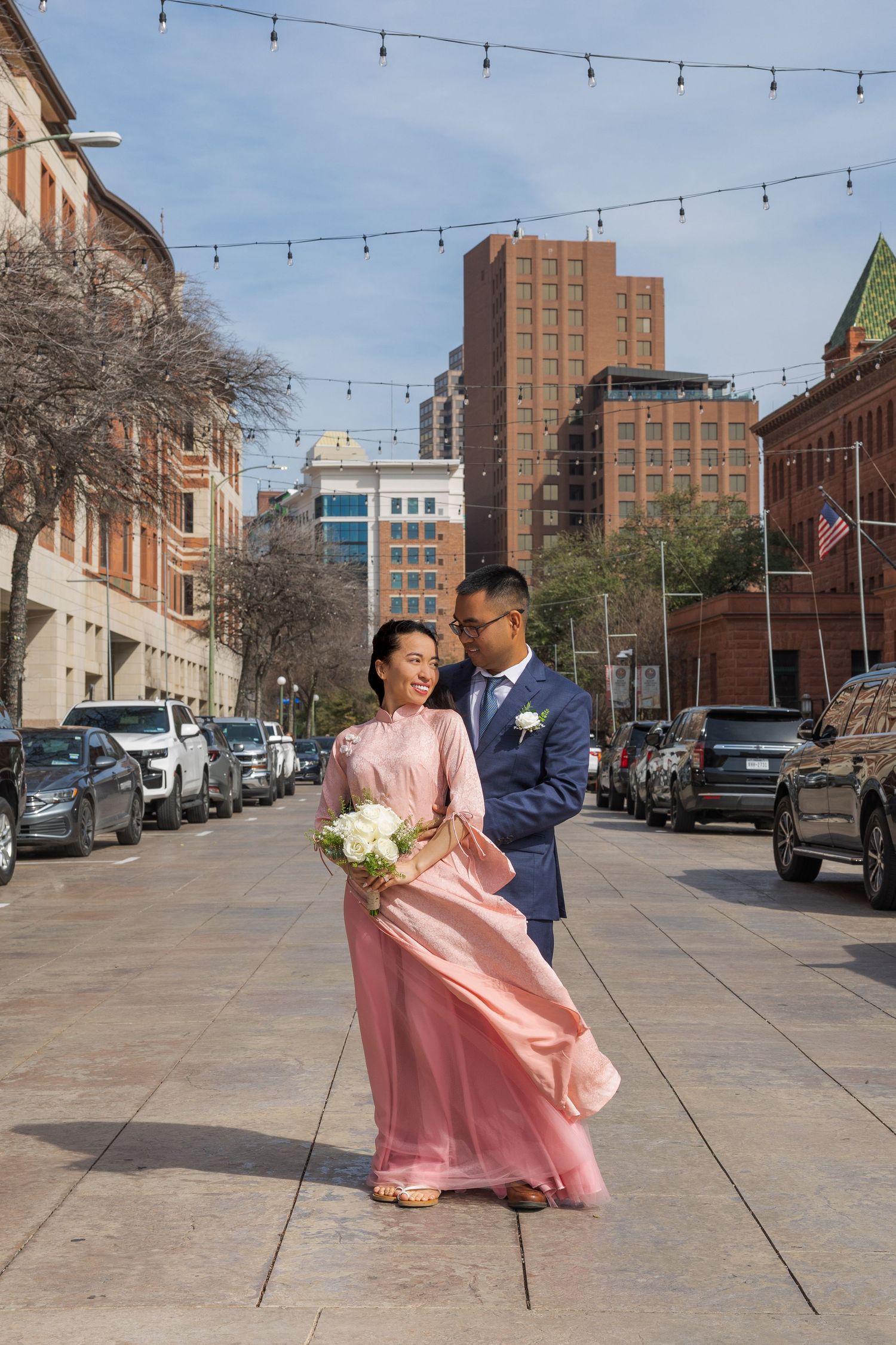 A couple in formal attire dances on a city street with string lights overhead and brick buildings in the background.