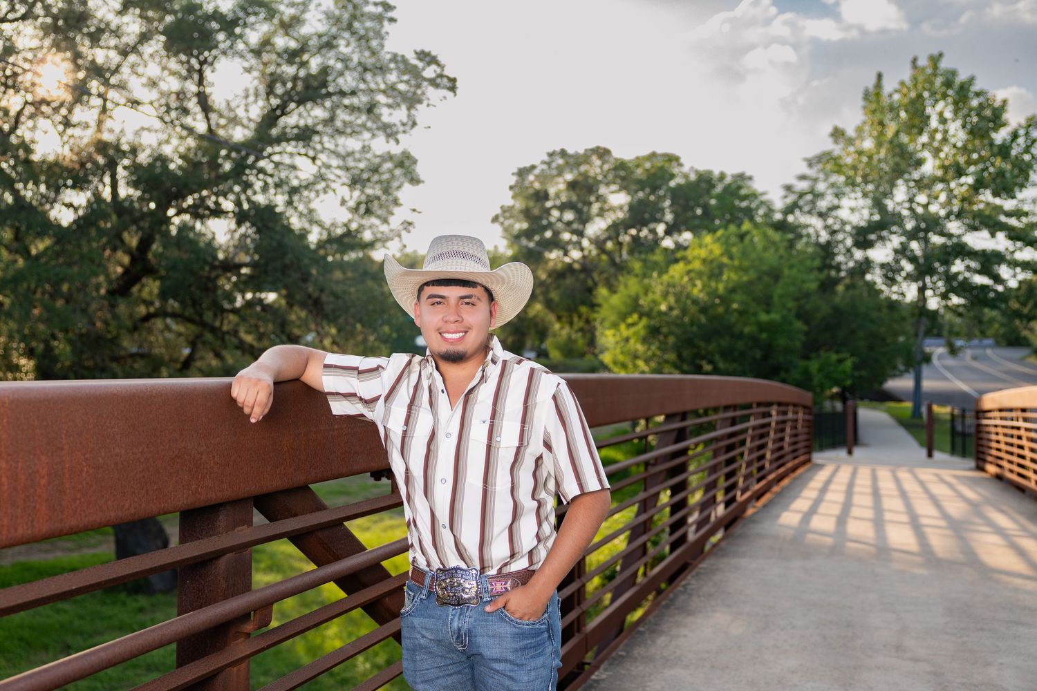 Young man in cowboy hat and striped shirt leaning on a rustic bridge railing outdoors.