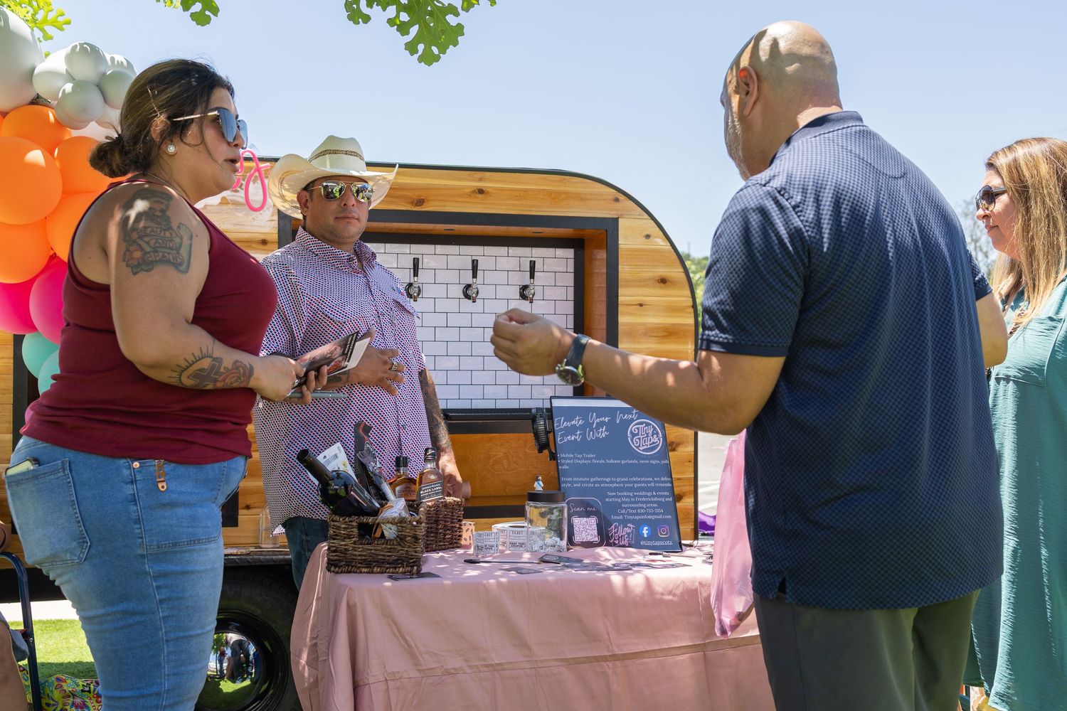 People gathered around an outdoor vendor booth with a small trailer at a sunny festival event.