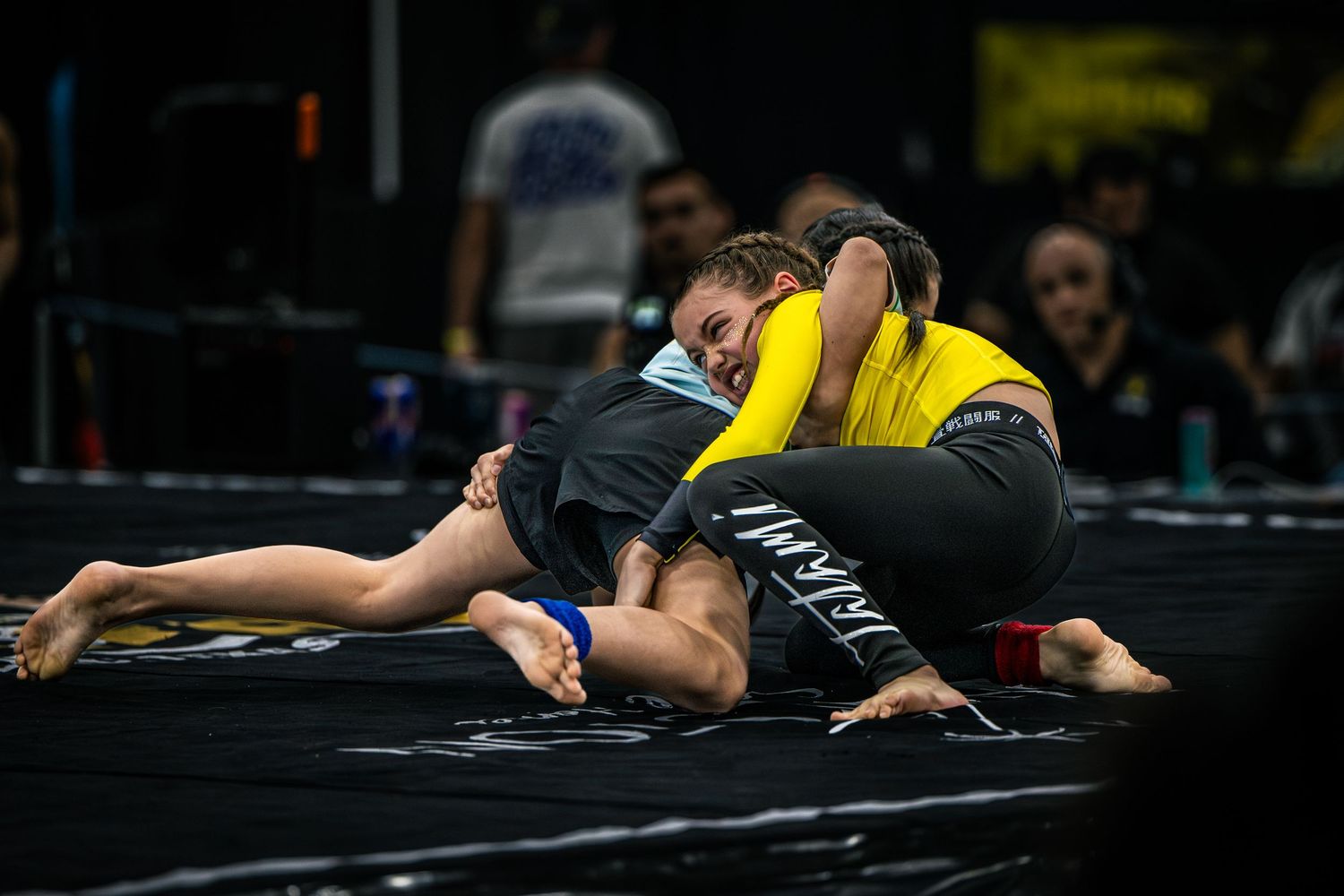 Two female grapplers compete in a BJJ match, one in yellow rashguard applying a submission hold on the mat.