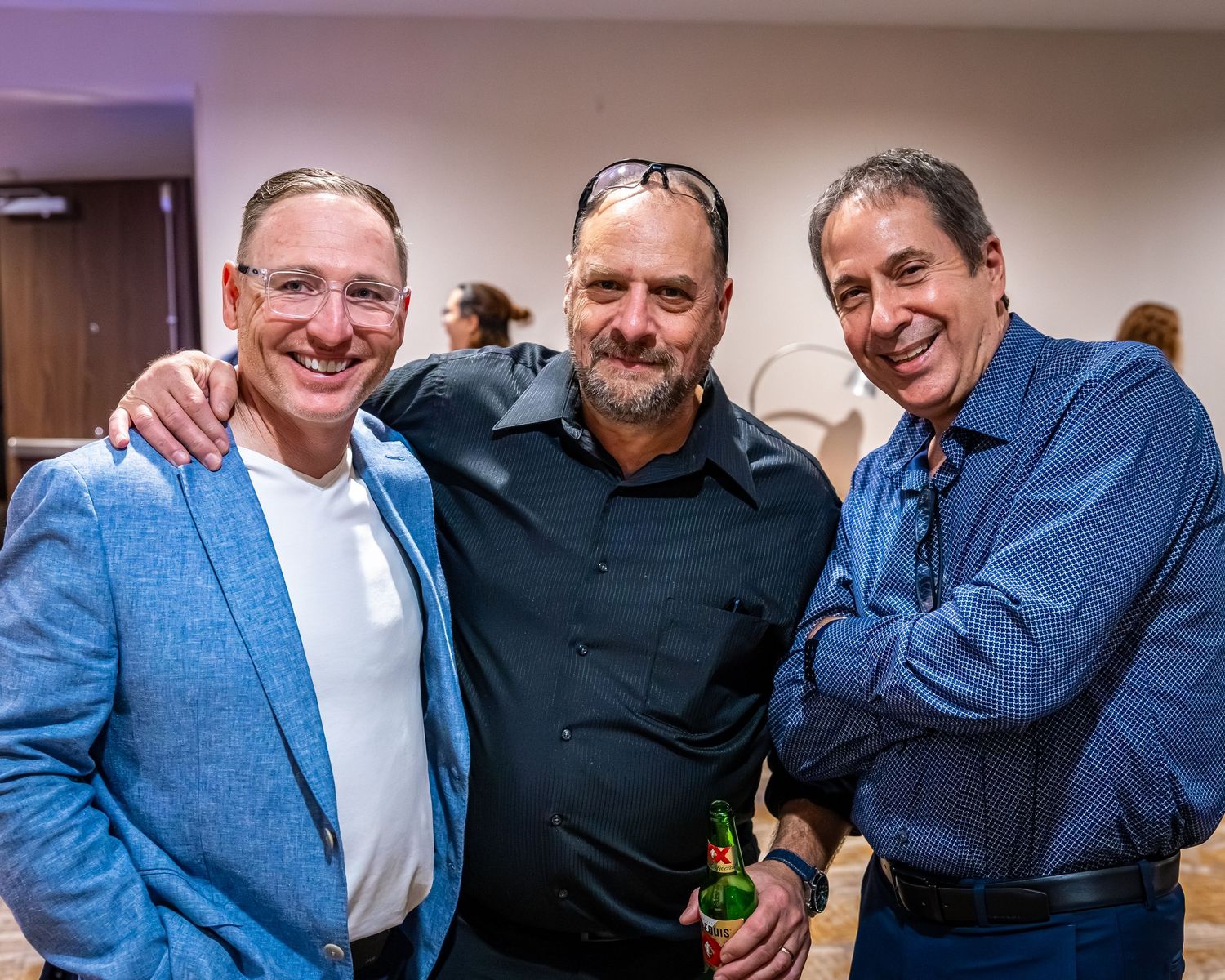 Three smiling men pose together at a social event, one in a blue blazer, one in black, one in a blue shirt.