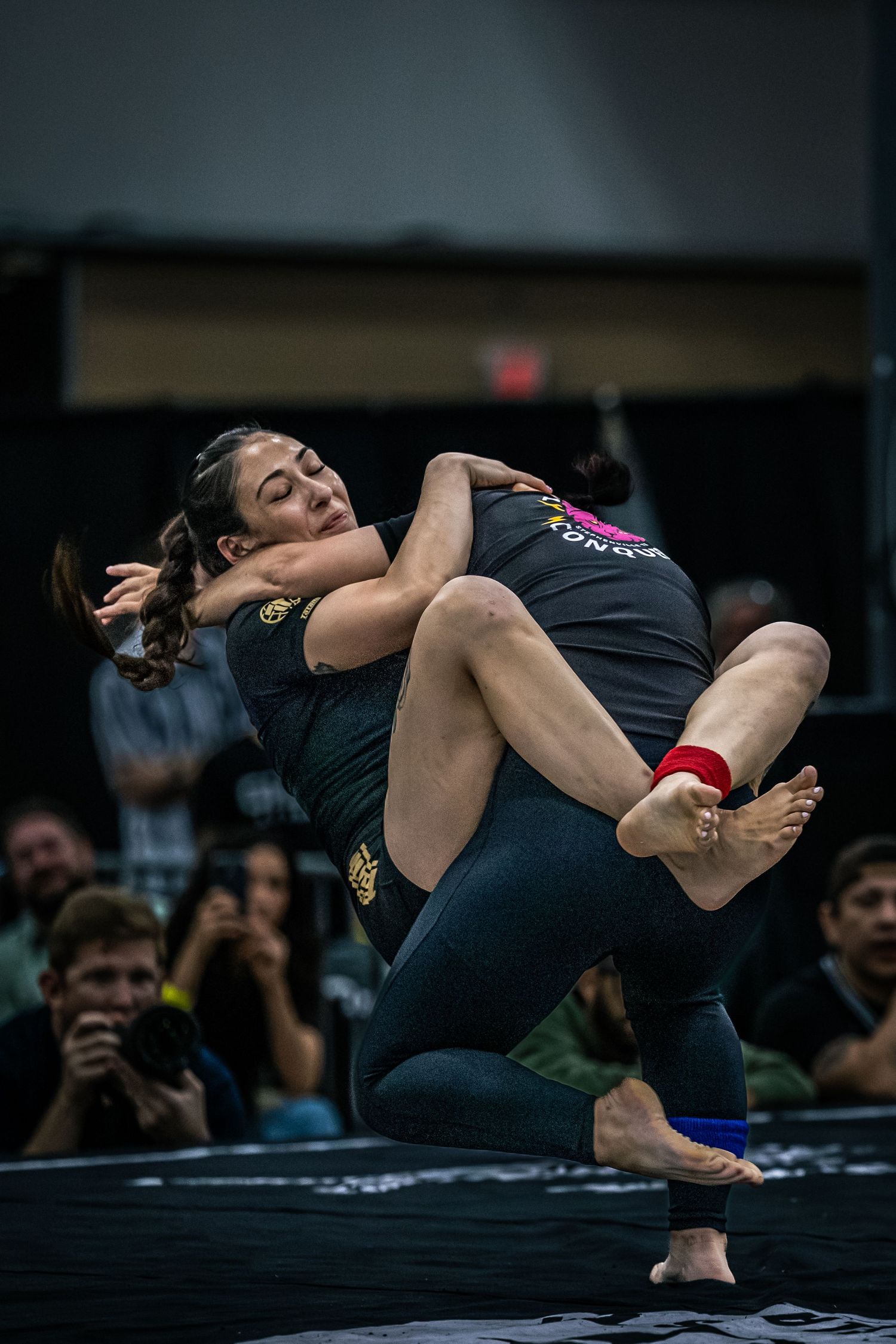 Two women competing in a grappling match at an indoor martial arts tournament with spectators watching.