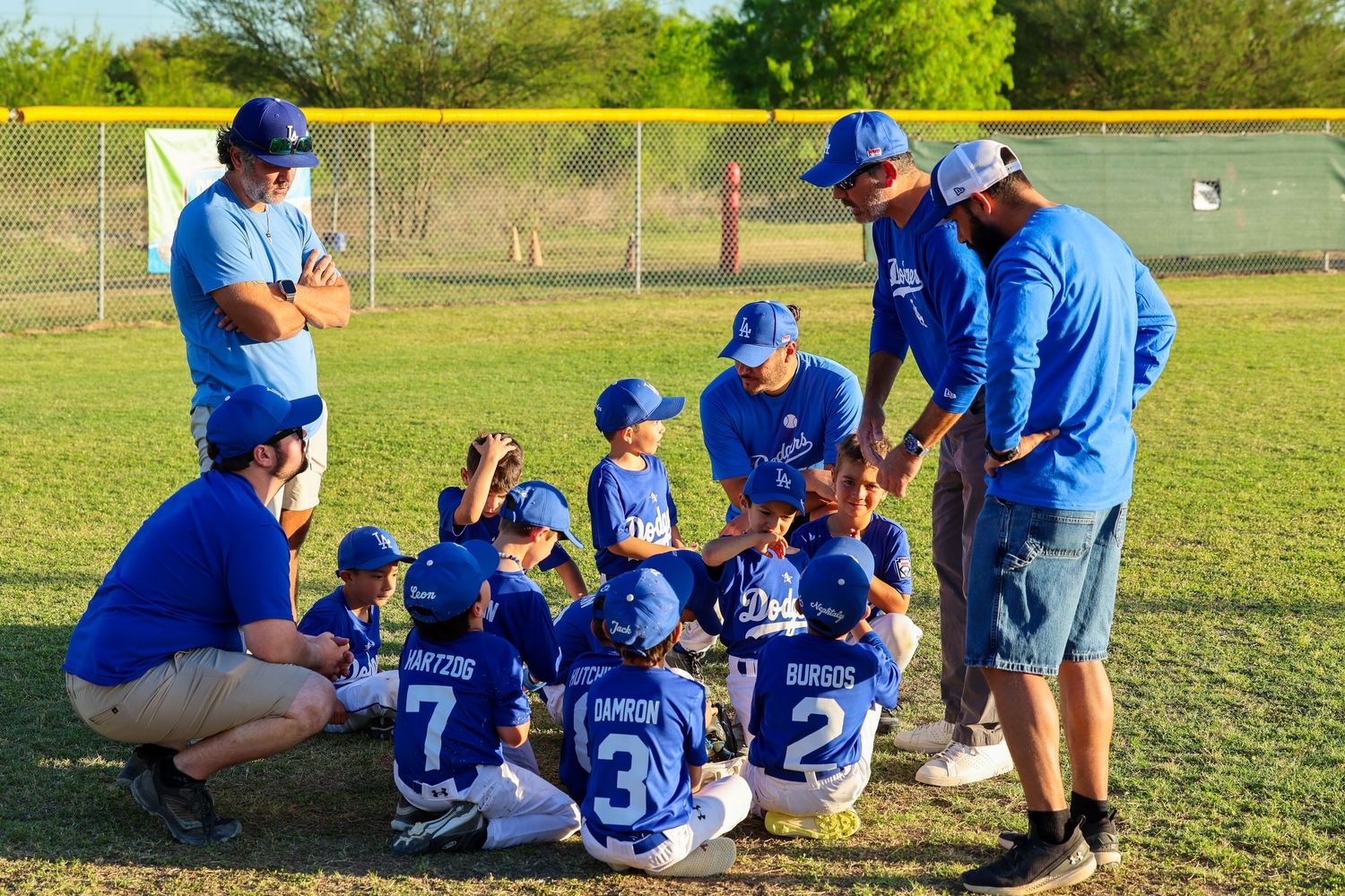 Youth baseball team in blue uniforms huddles with coaches on a green field during a little league game.