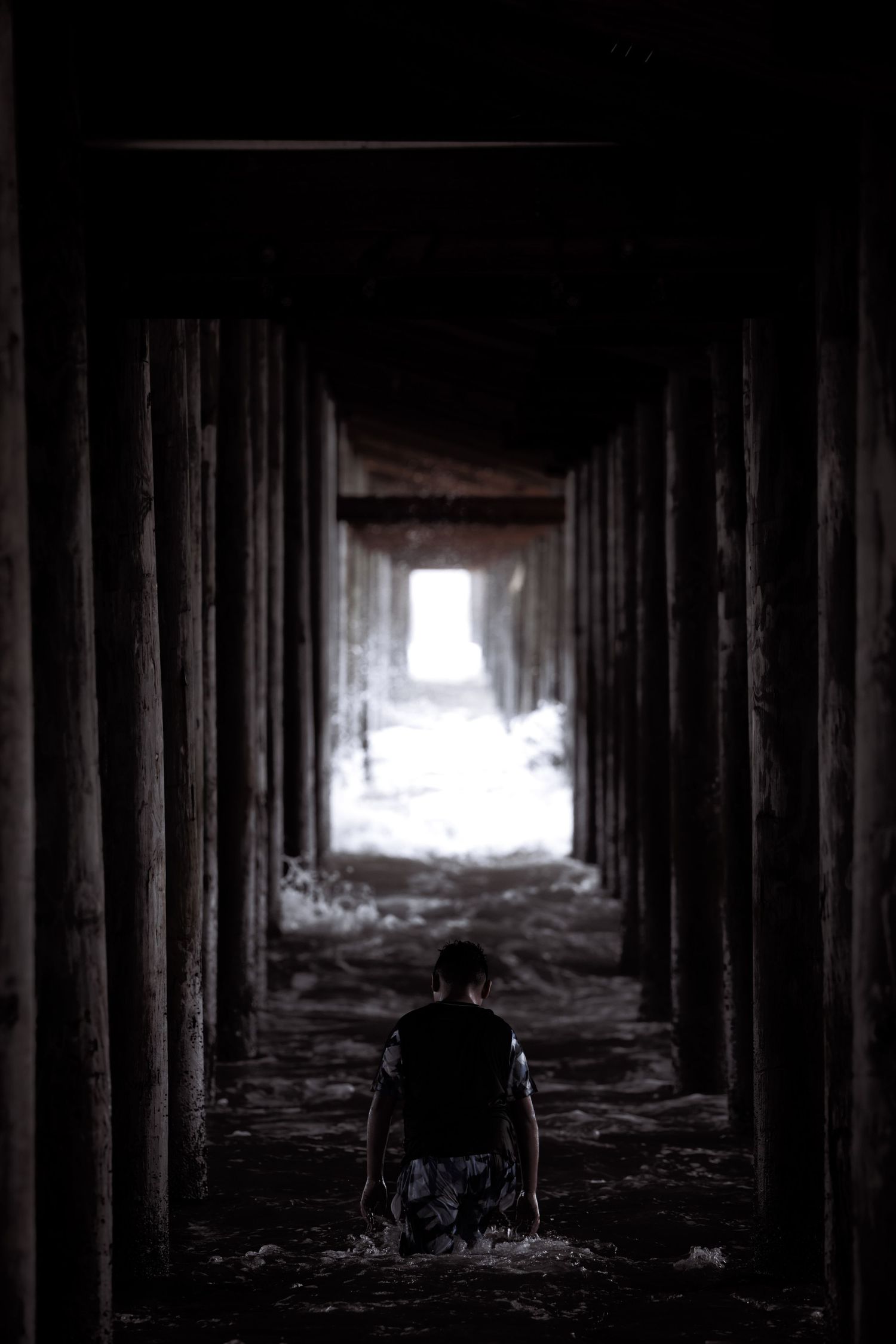 Person wading through dark tunnel under pier with light visible at the end, creating dramatic contrast.