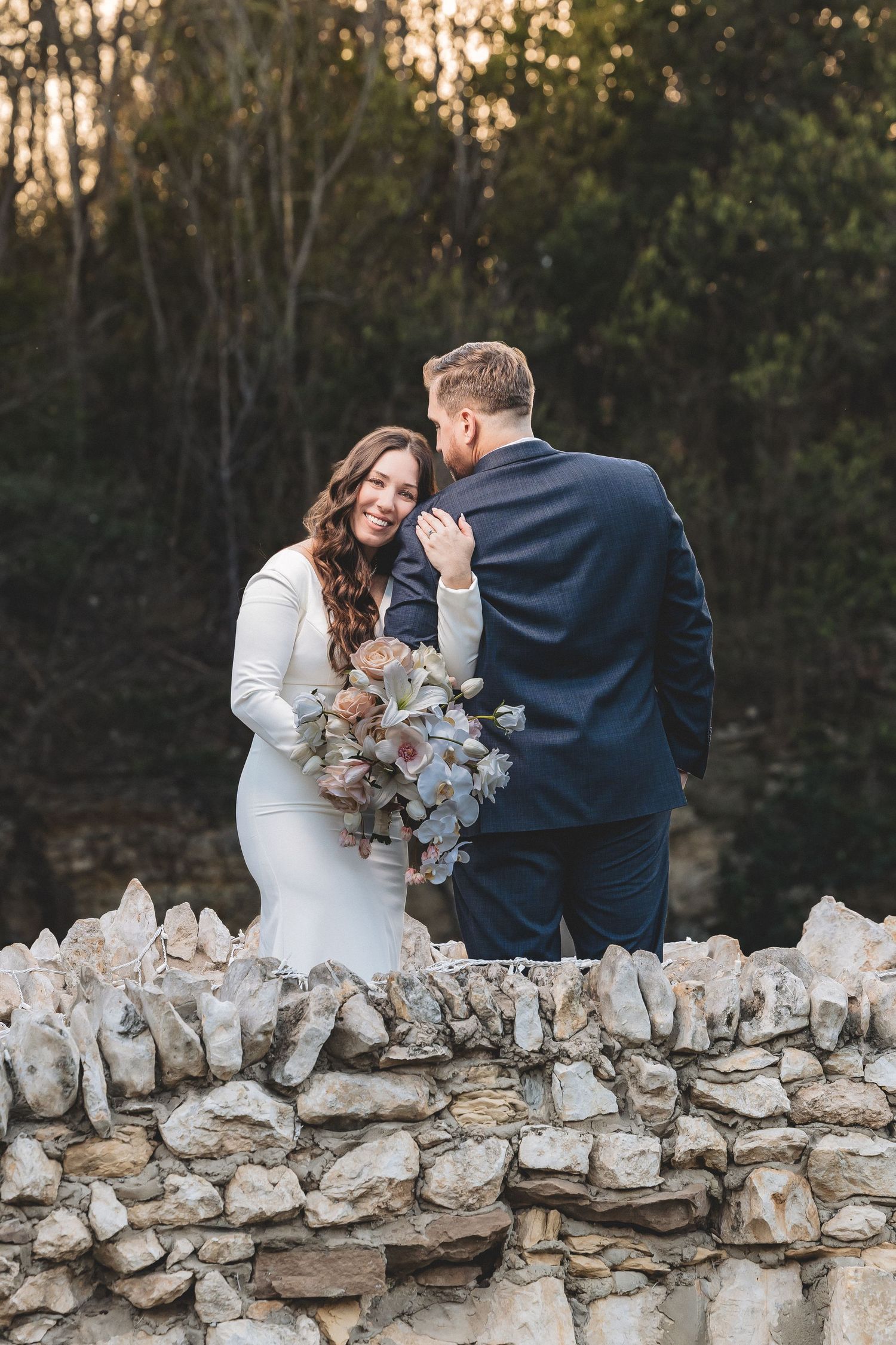 A romantic couple in wedding attire share an intimate moment behind a rustic stone wall at sunset.
