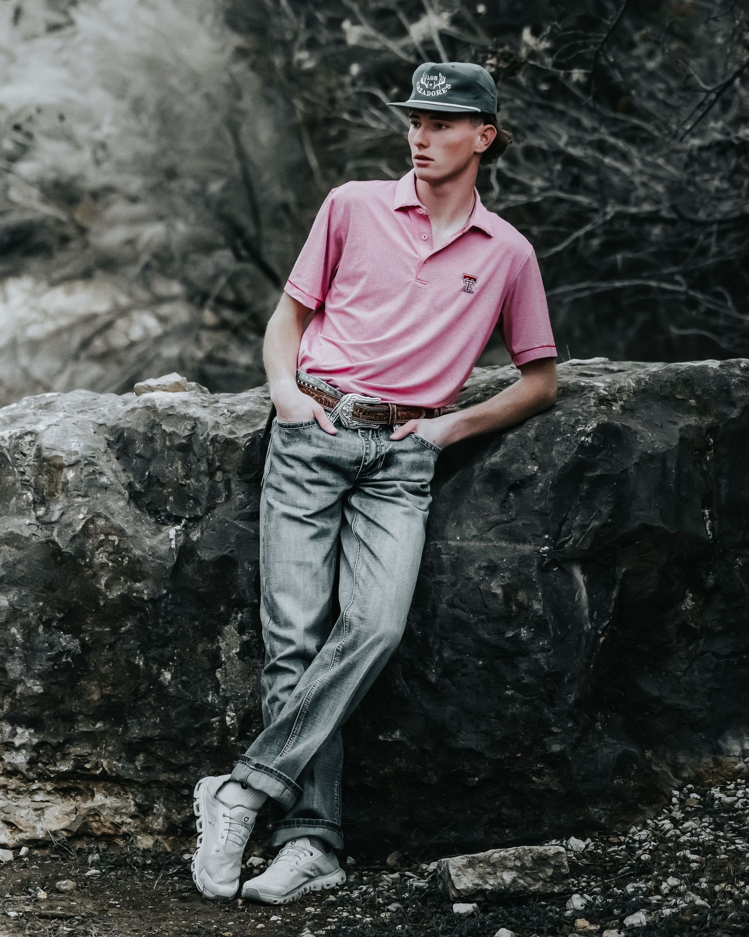 Person in a pink polo shirt and light jeans sits casually on a rocky surface in a moody outdoor setting.