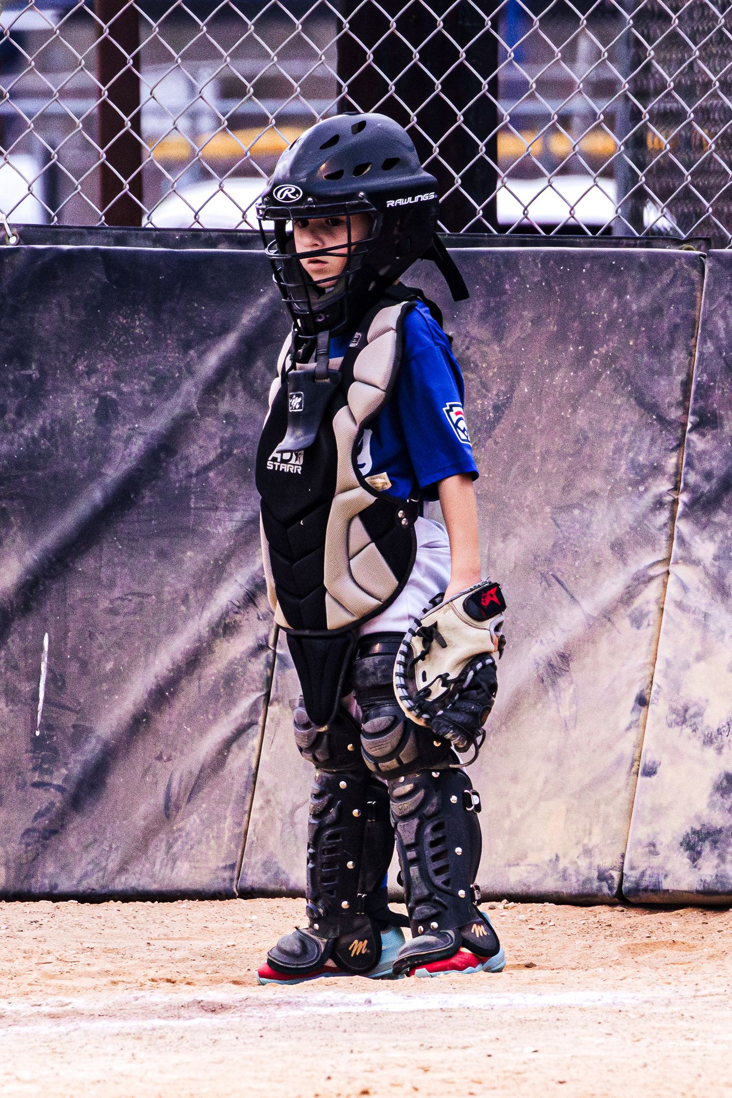 Young baseball catcher in full protective gear including helmet, chest protector, and shin guards standing on a dirt field.