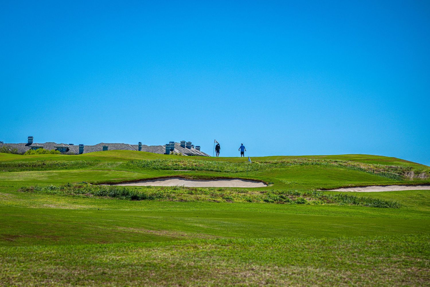 Golfers on a lush green golf course with sand bunkers under a clear blue sky.