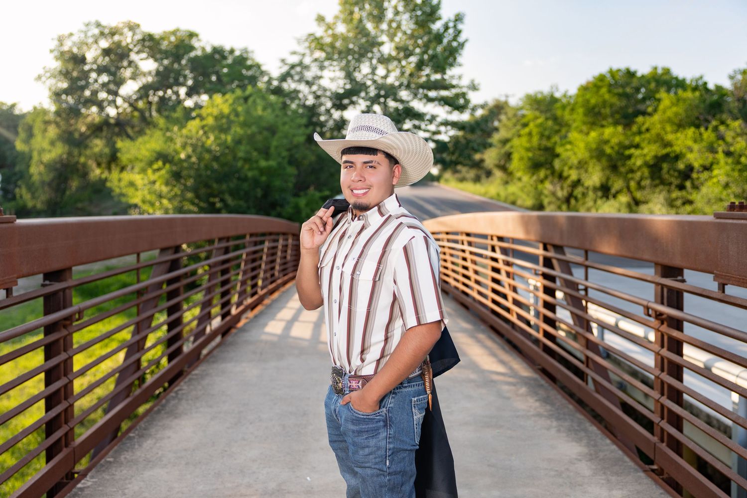 Young man in cowboy hat and striped shirt smiling on a bridge surrounded by lush green trees.