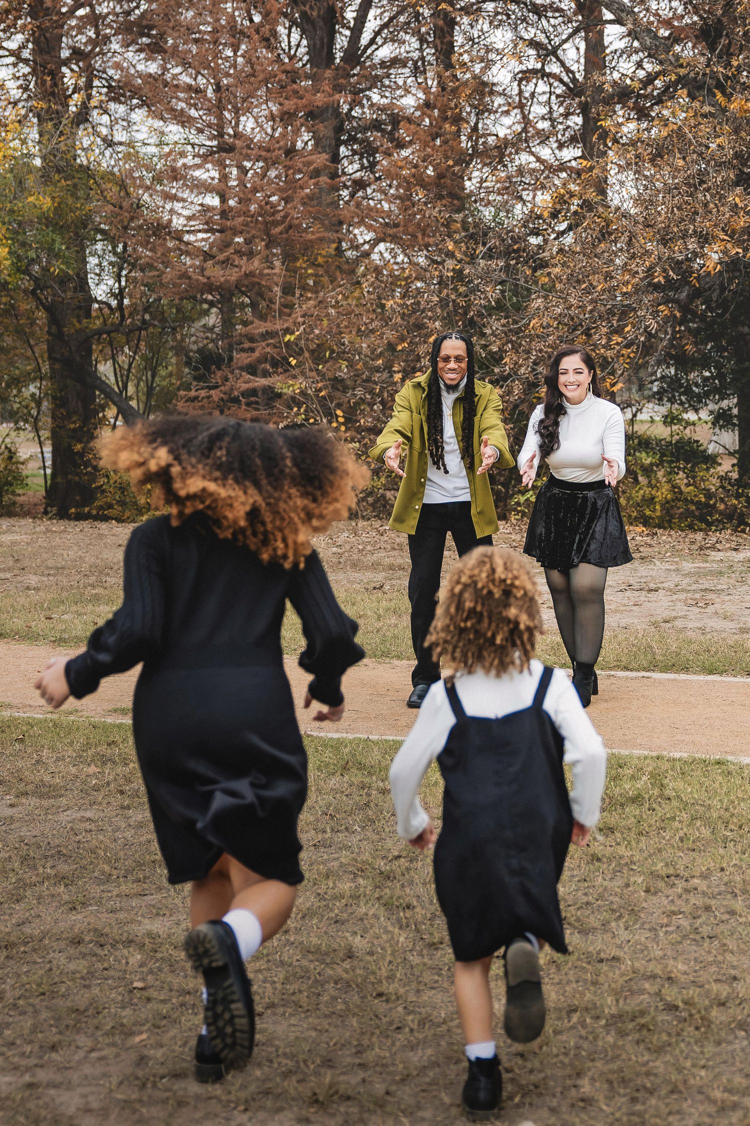 Two children in dark dresses run towards two others standing in an autumn park setting with brown trees in background.