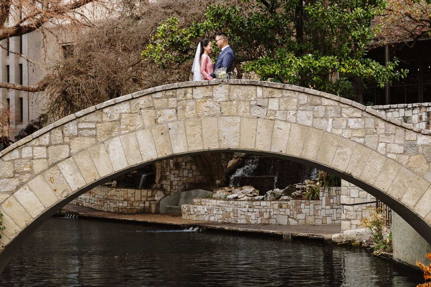 Stone bridge reflects in calm water surrounded by lush greenery and trees in tranquil setting.