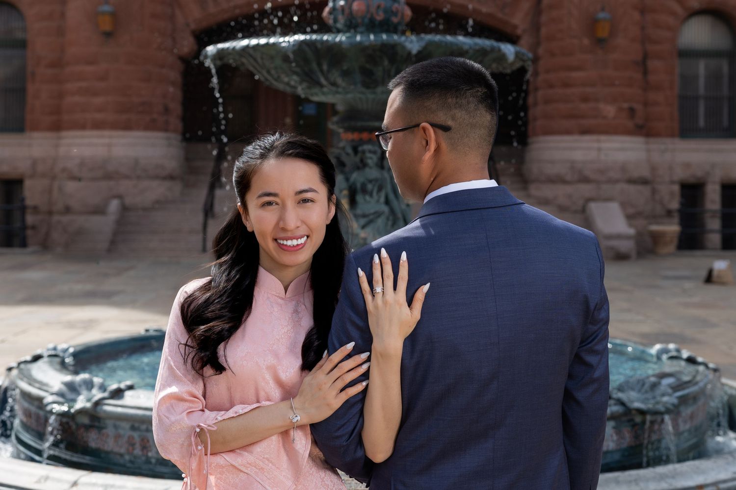 A couple in formal attire embraces near a brick building with a fountain in the background.