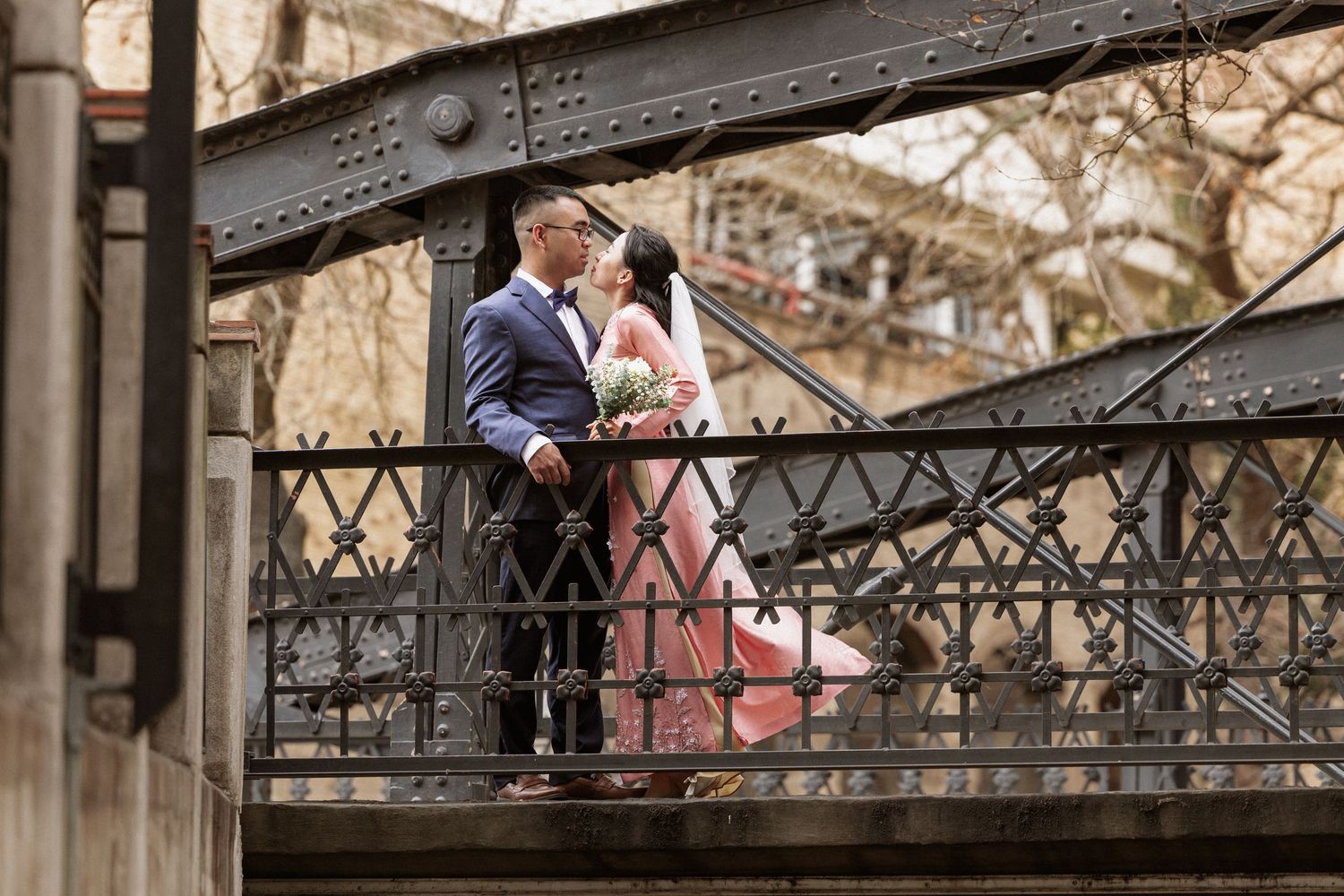 Couple sharing a romantic moment on an ornate iron bridge, bride in pink dress holding bouquet, groom in navy suit.