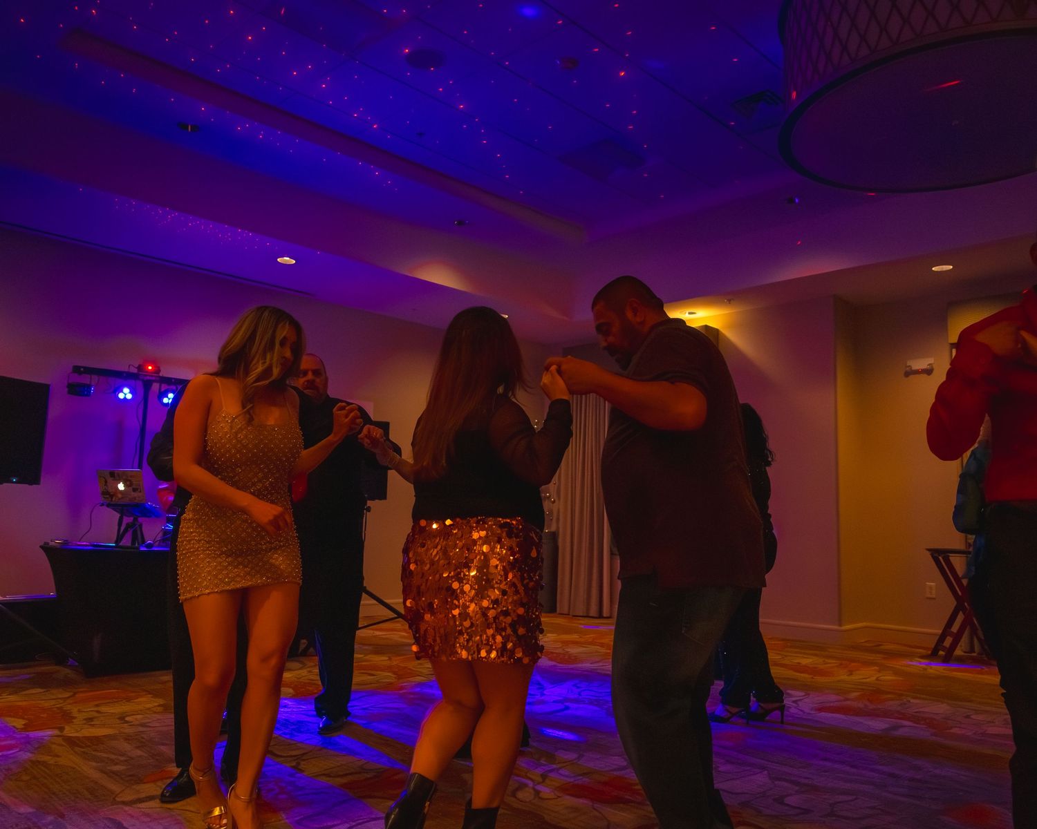 People dancing at an indoor party with blue and purple lighting, wearing festive attire including sequin skirts.