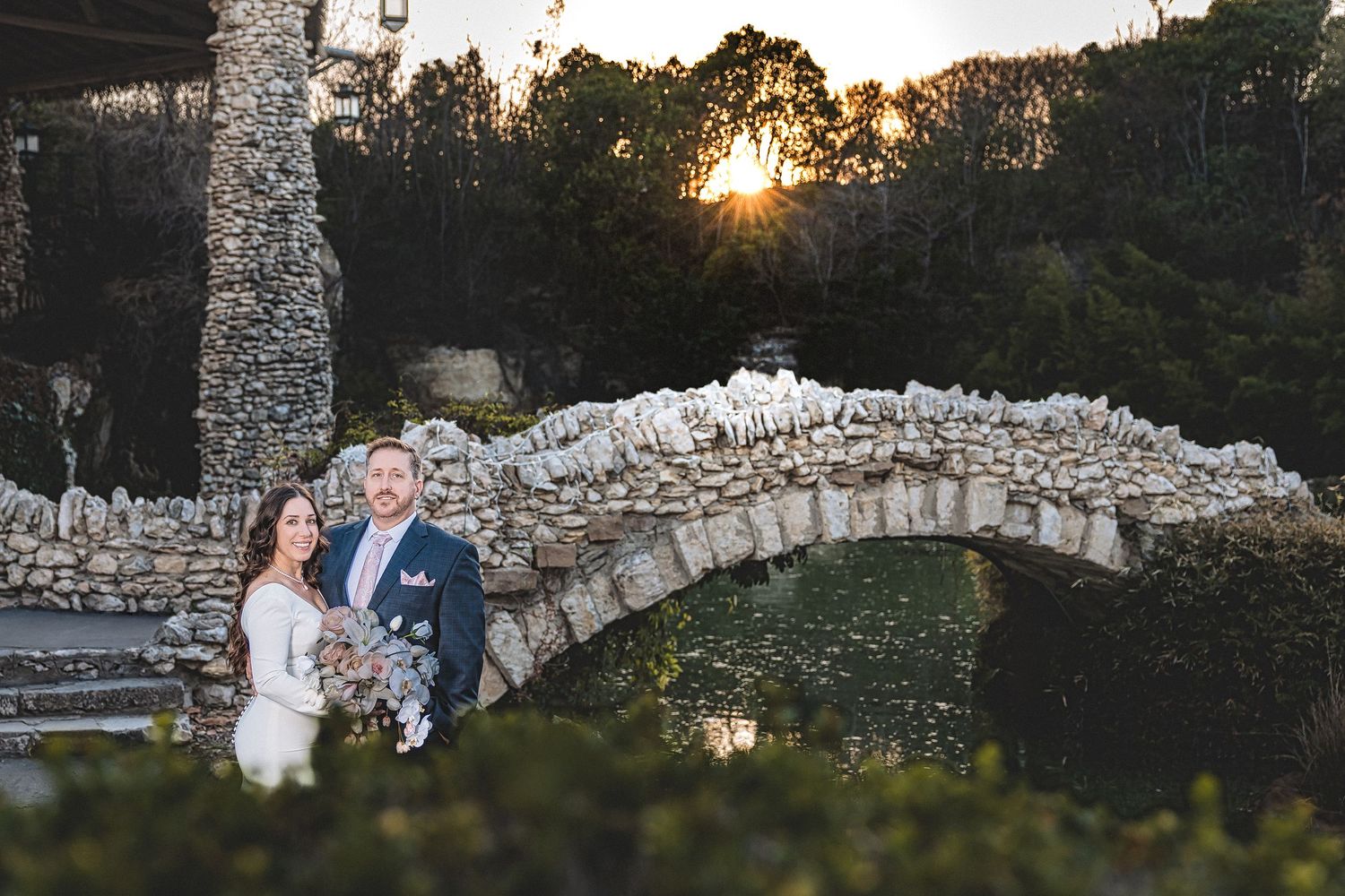 A couple shares a romantic moment at sunset near a stone wall in a scenic outdoor setting.