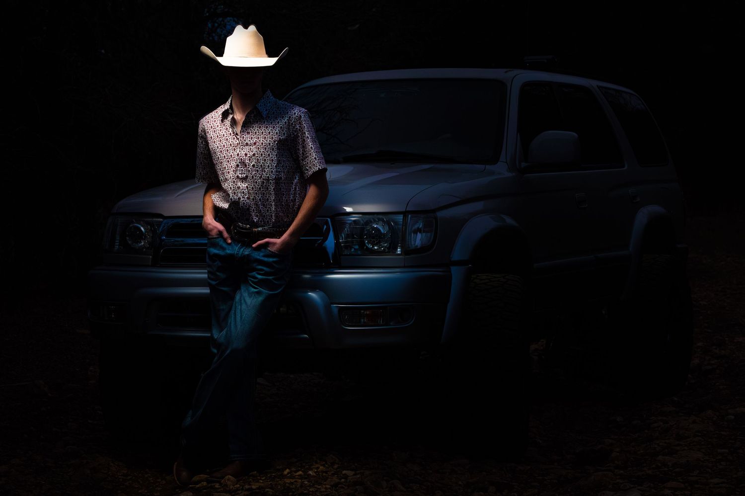 Man in white cowboy hat leans against silver SUV truck at night in dramatic dark lighting.