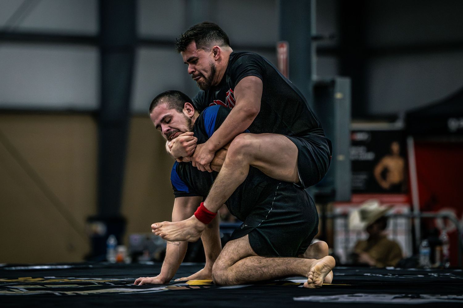 Two MMA fighters grappling on a black mat during a training session in a gym, practicing submission wrestling techniques.