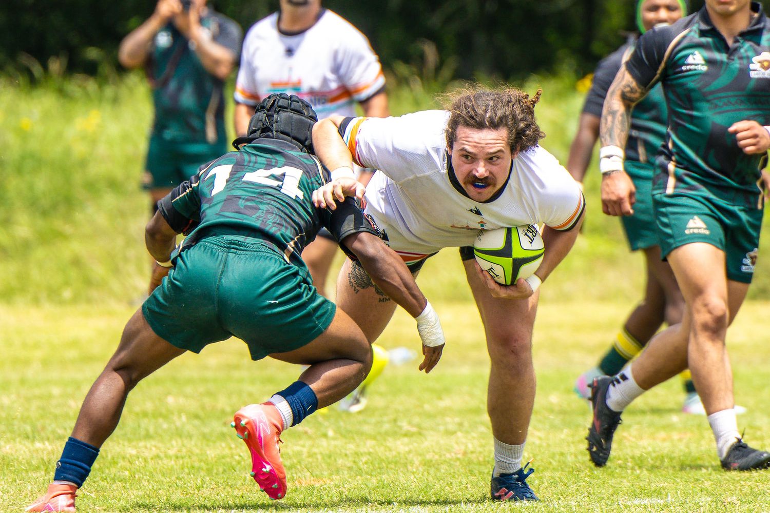 Rugby player in white jersey dives forward with ball while tackled by opponent in green uniform outdoors.