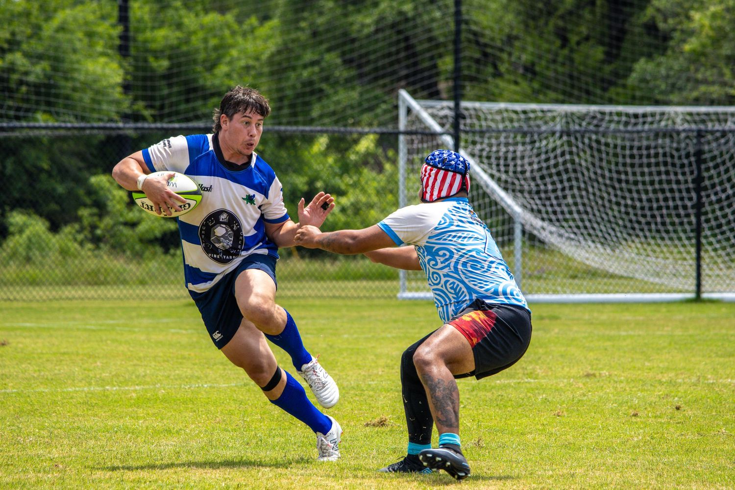 Two rugby players compete on a green field, one carrying the ball while evading a tackle during an outdoor match.