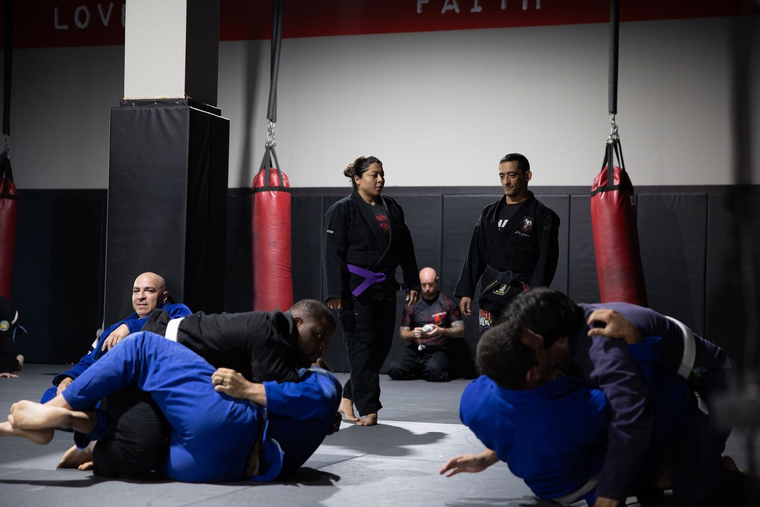 Martial arts instructor in purple belt observing BJJ students practicing grappling techniques on gym mats.