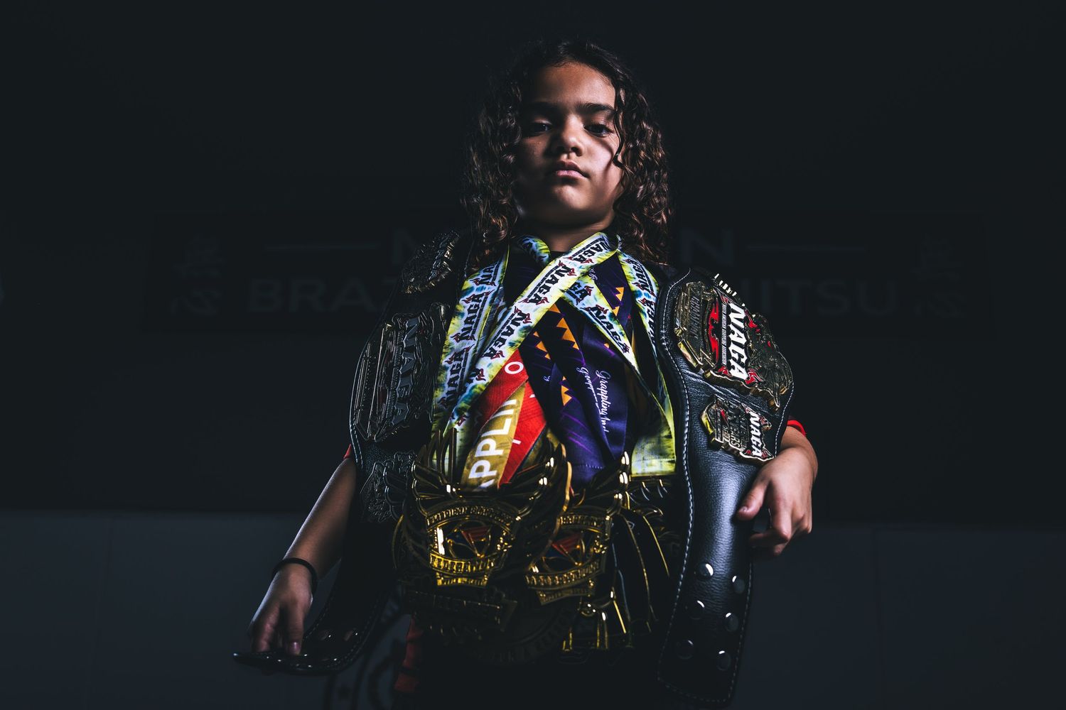 Young female fighter wearing multiple championship belts poses confidently in dramatic dark lighting.