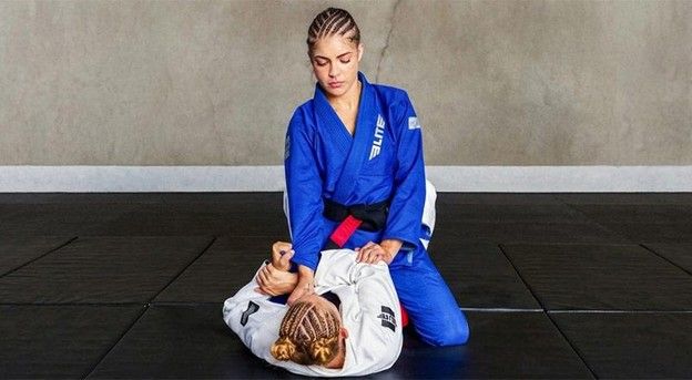 Woman in blue BJJ gi practicing ground grappling technique on black martial arts mat.
