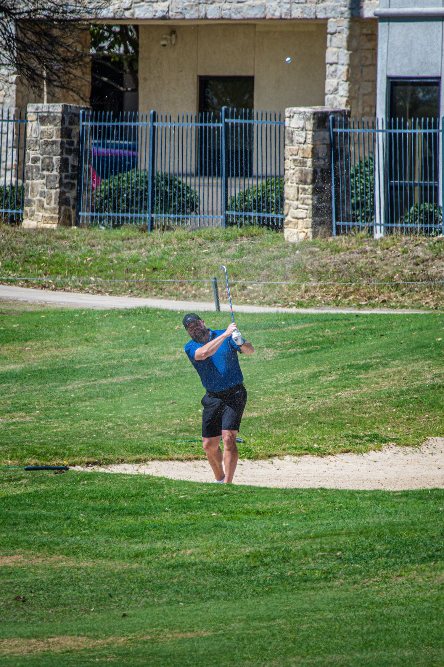 Golfer in blue shirt hits shot from sand bunker on sunny golf course near stone building.