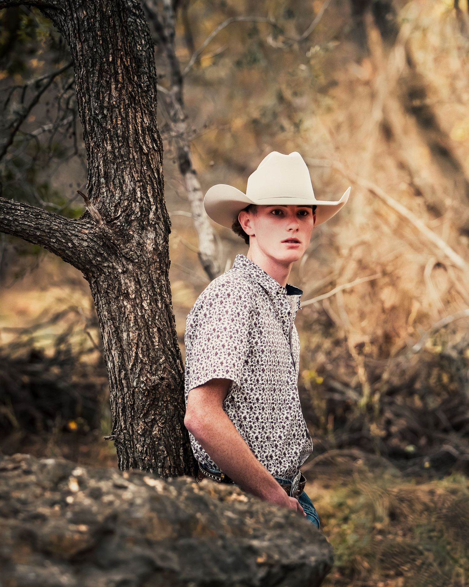 A person in a white cowboy hat and patterned shirt poses thoughtfully against a wooded outdoor backdrop.