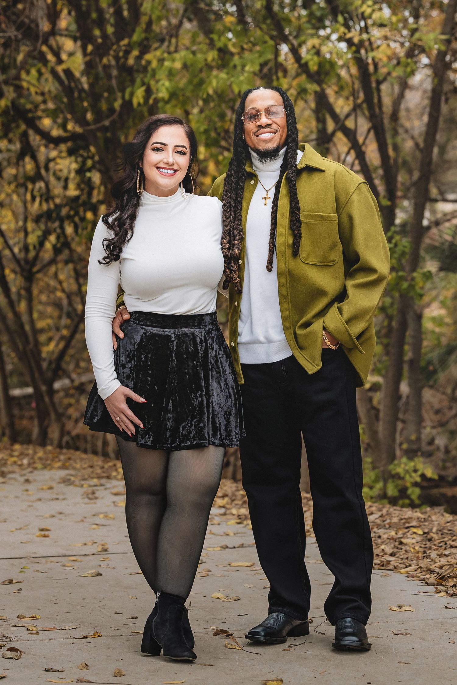 A couple in formal attire poses together on a path covered in autumn leaves, wearing white tops and dark bottoms.