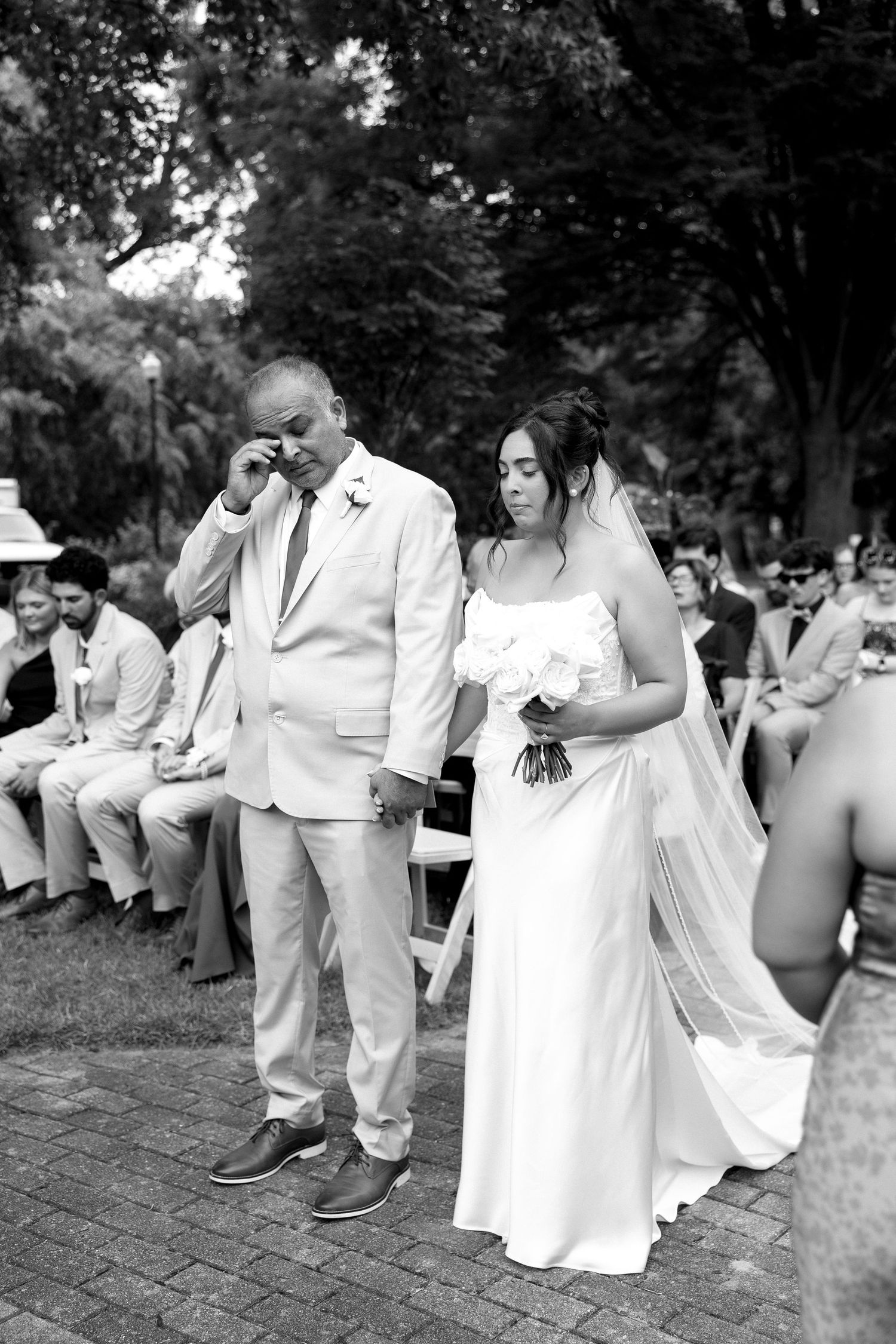 Emotional father wipes tears while walking bride down outdoor wedding aisle in black and white photo.