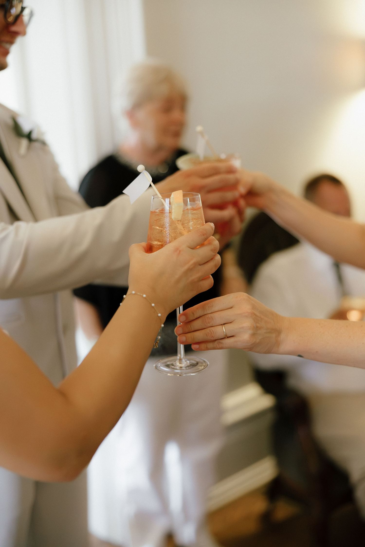 Wedding guests clink champagne glasses in celebratory toast, bride wearing pearl bracelet and engagement ring.
