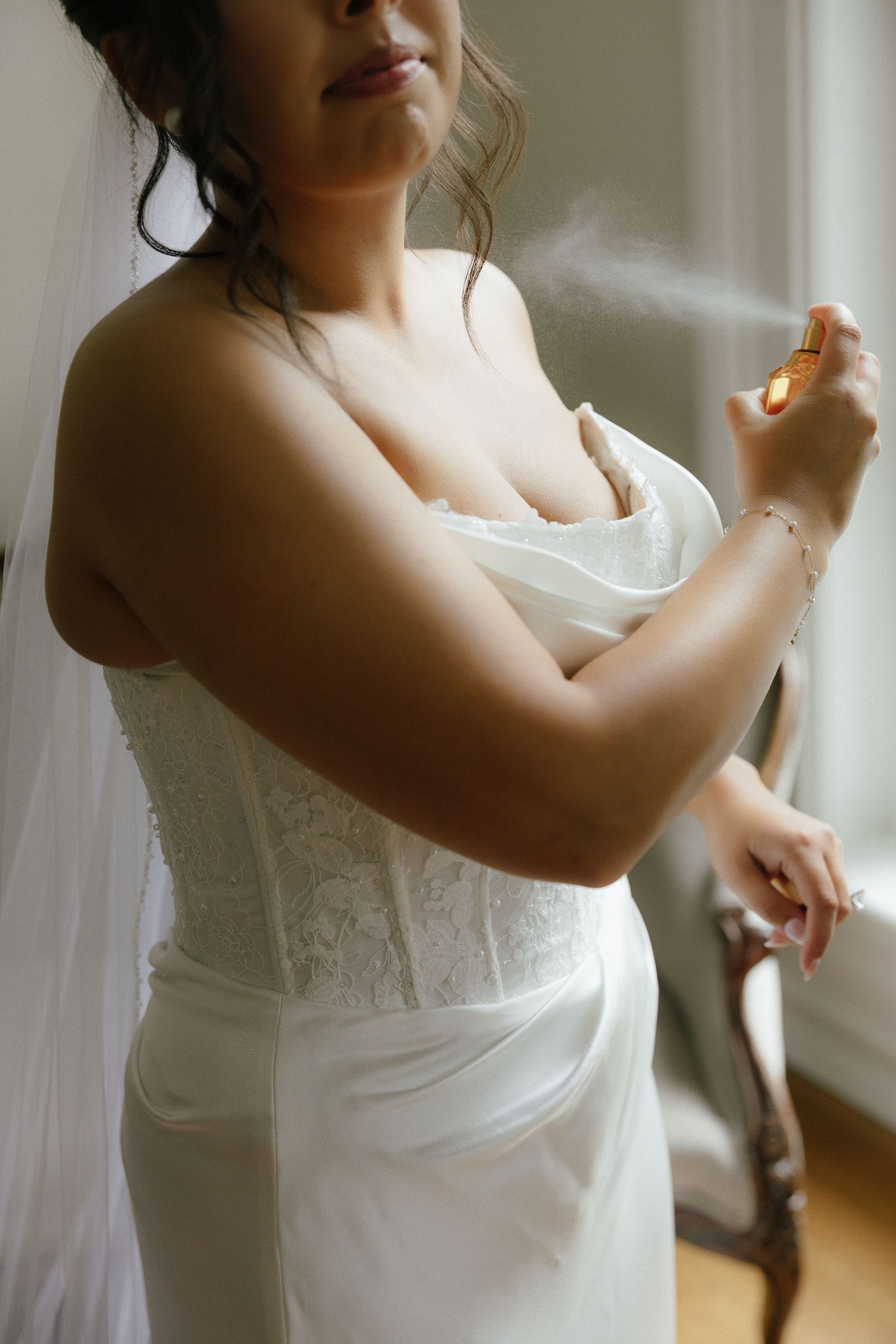 Bride in white lace wedding dress applying perfume while getting ready near a window.