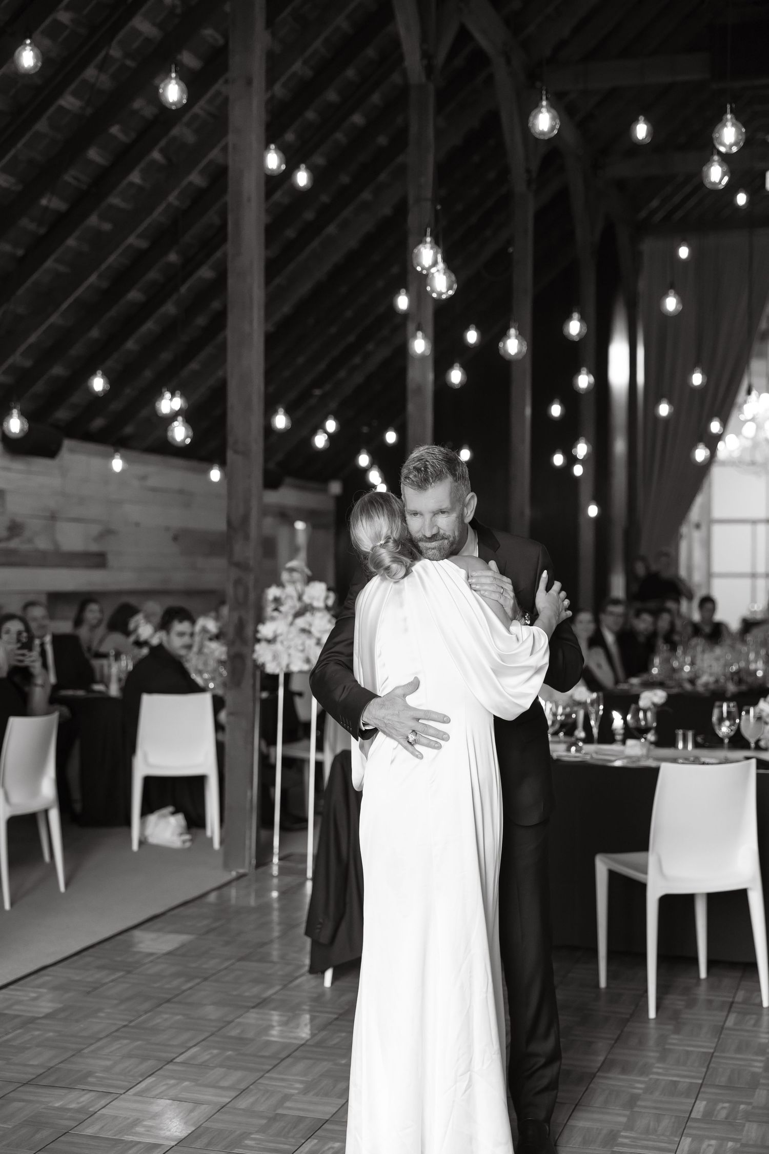Black and white photo of bride and groom sharing first dance under Edison bulb lights in barn.