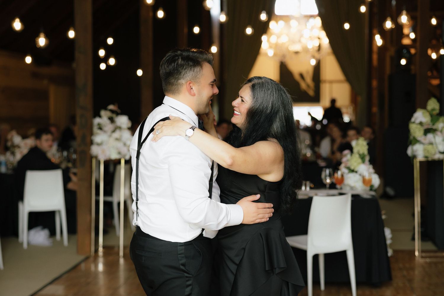 Groom and mother sharing emotional dance at wedding reception with warm string lights.