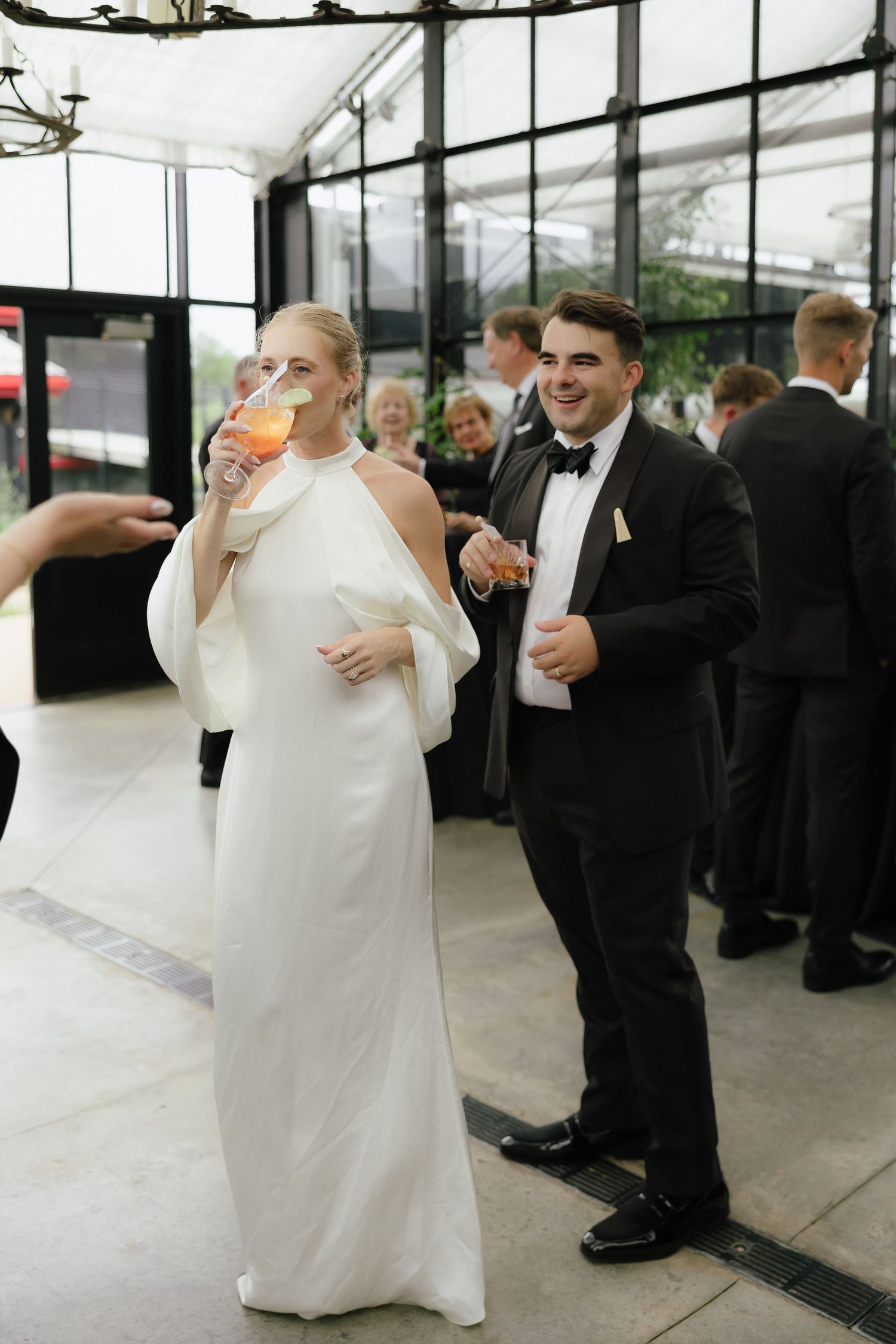 Smiling bride in white cape gown sipping cocktail beside groom in tuxedo at wedding reception venue.