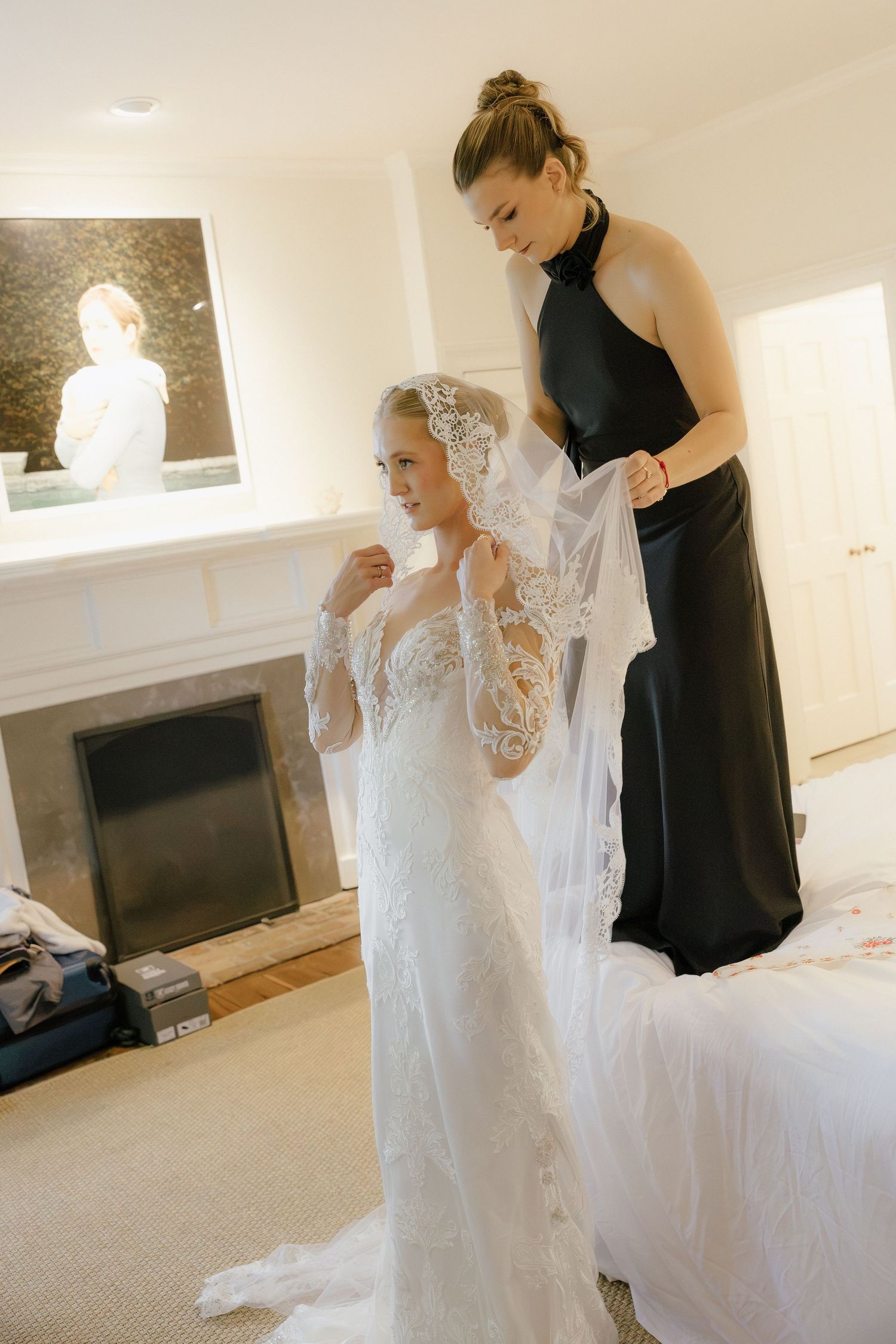 Bridesmaid in black dress helping blonde bride arrange lace veil in elegant bridal suite.