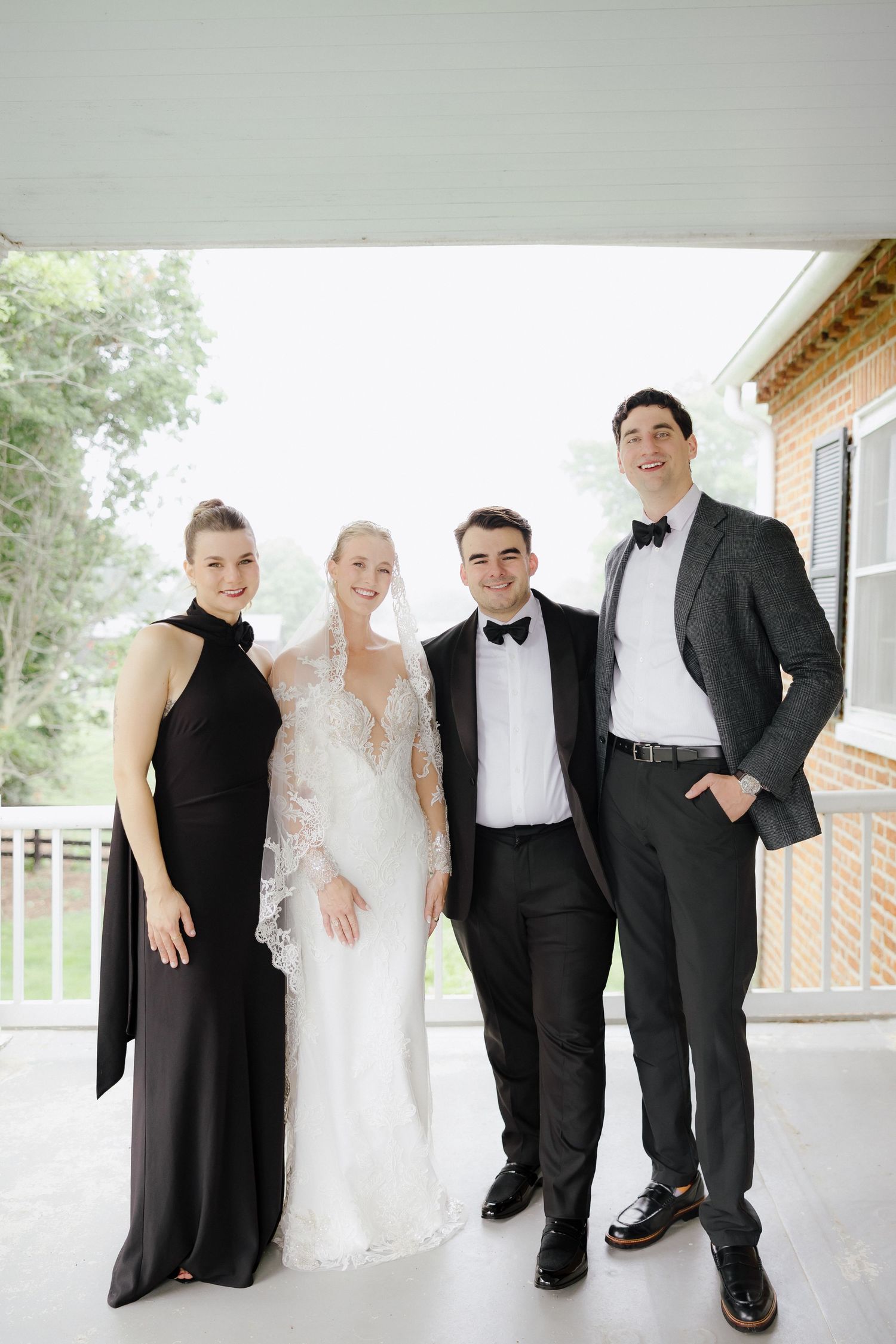 Wedding party of four posing on porch, bride in lace gown with veil, groom in tuxedo, bridesmaid in black dress.