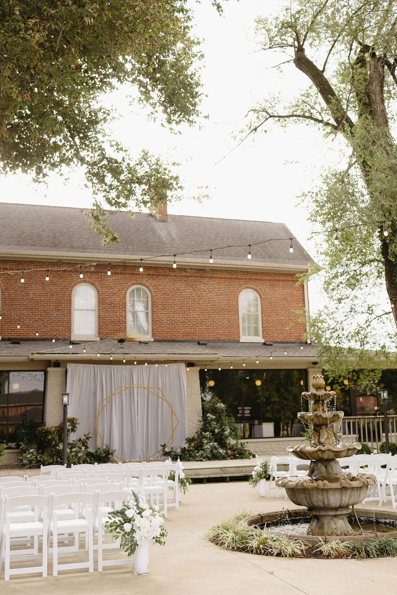 Outdoor wedding ceremony setup with white chairs and draping at a brick venue with string lights and fountain.