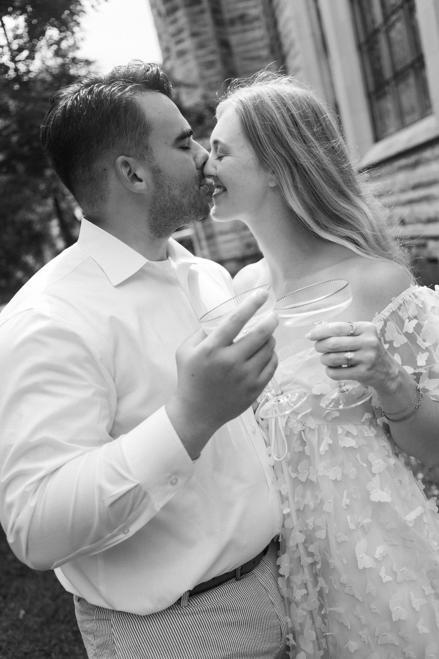 Black and white photo of a couple sharing a romantic kiss while holding champagne glasses outdoors near a stone building.