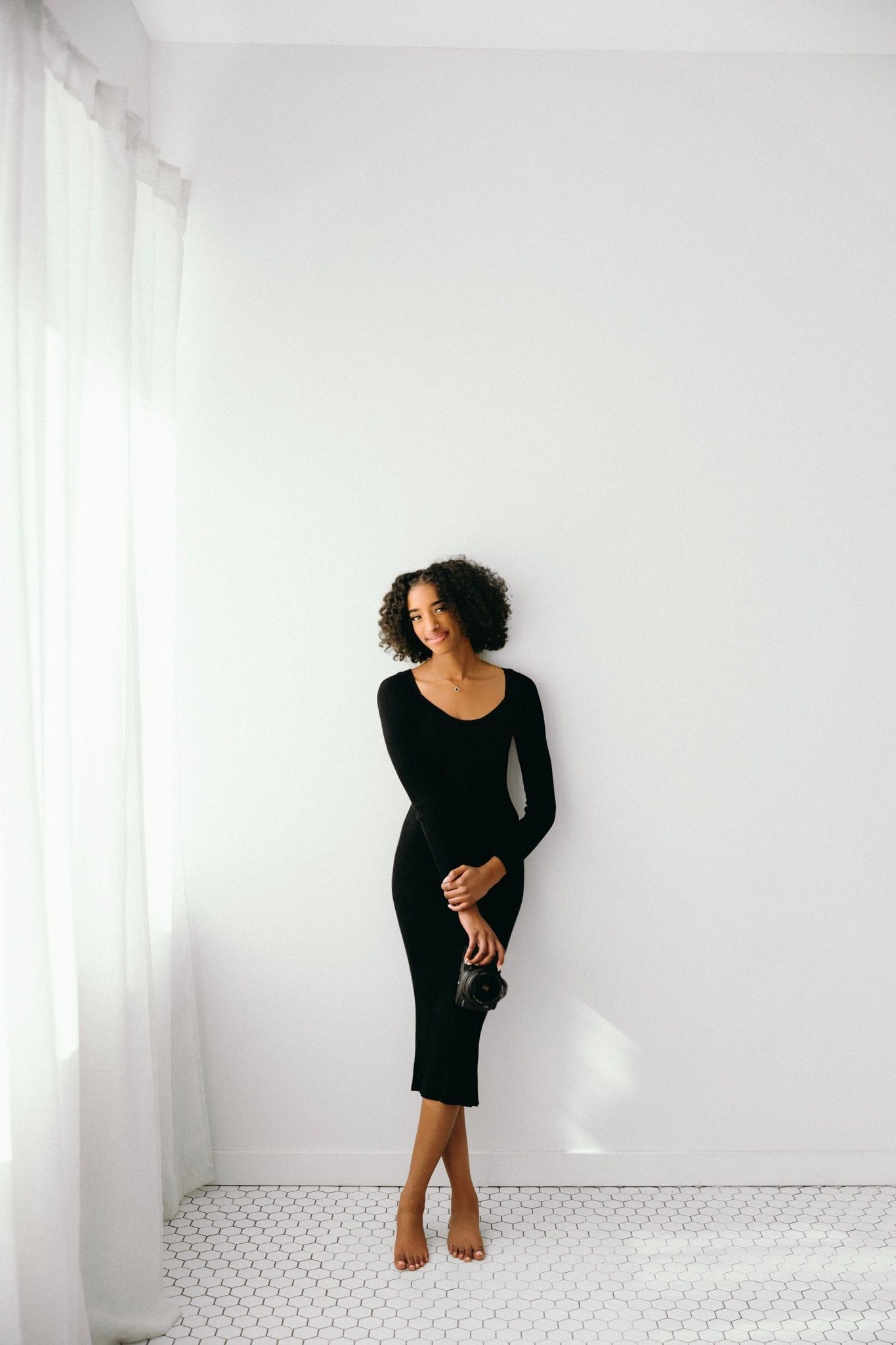 Woman in black long-sleeve midi dress and heels posing against white wall, holding clutch bag.